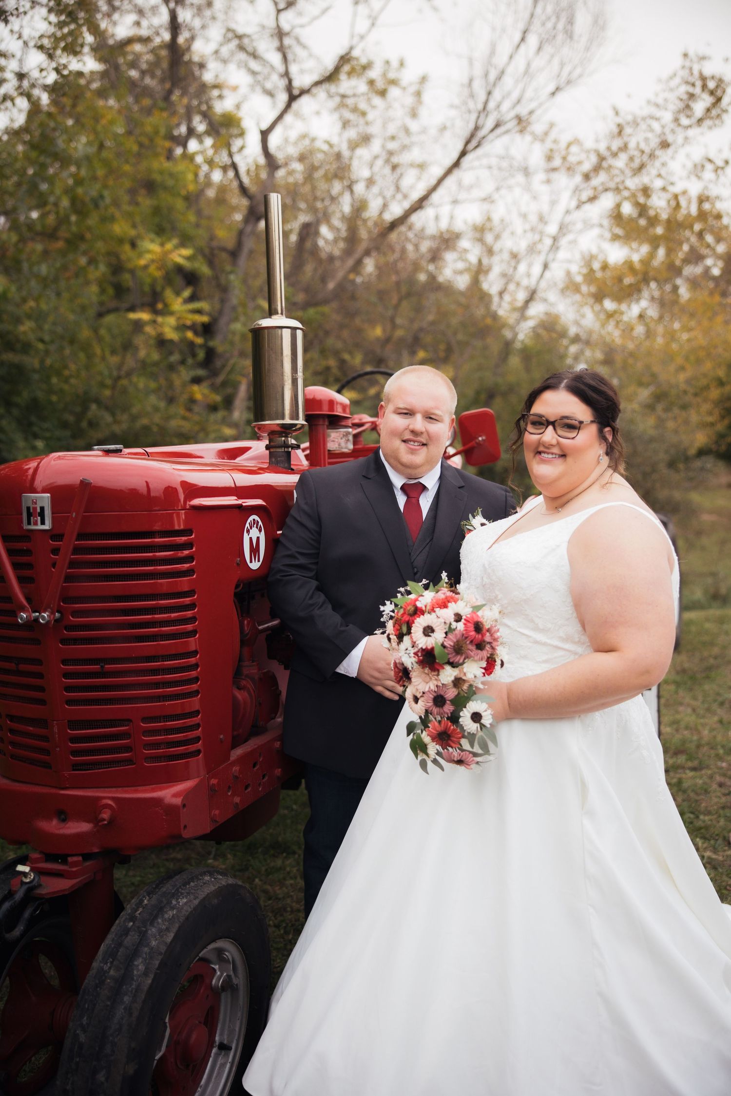 Wedding couple poses next to a vintage red farm tractor in an autumn outdoor setting.