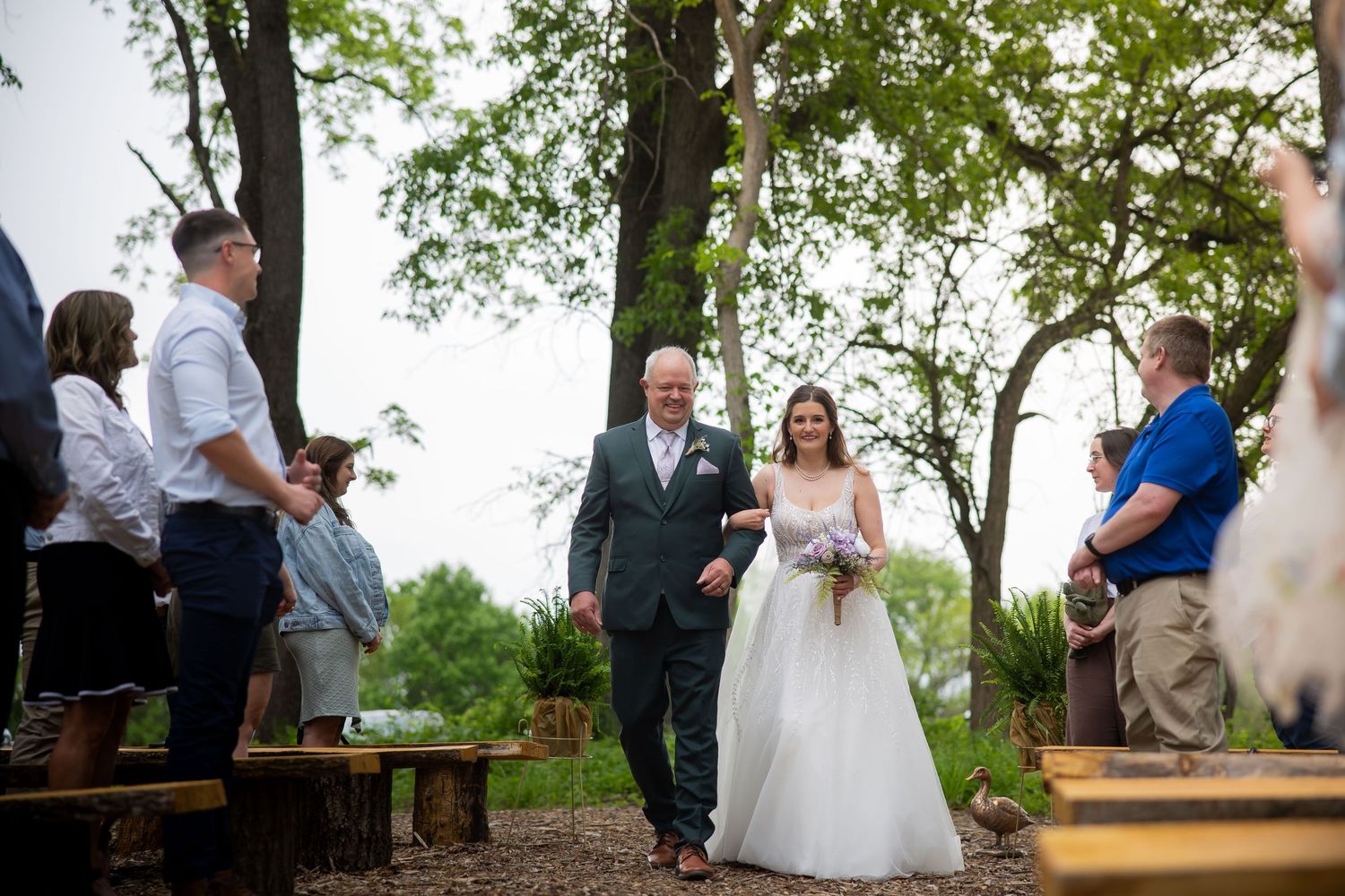 Bride in white gown and veil walks down outdoor woodland aisle with older gentleman at wedding ceremony.