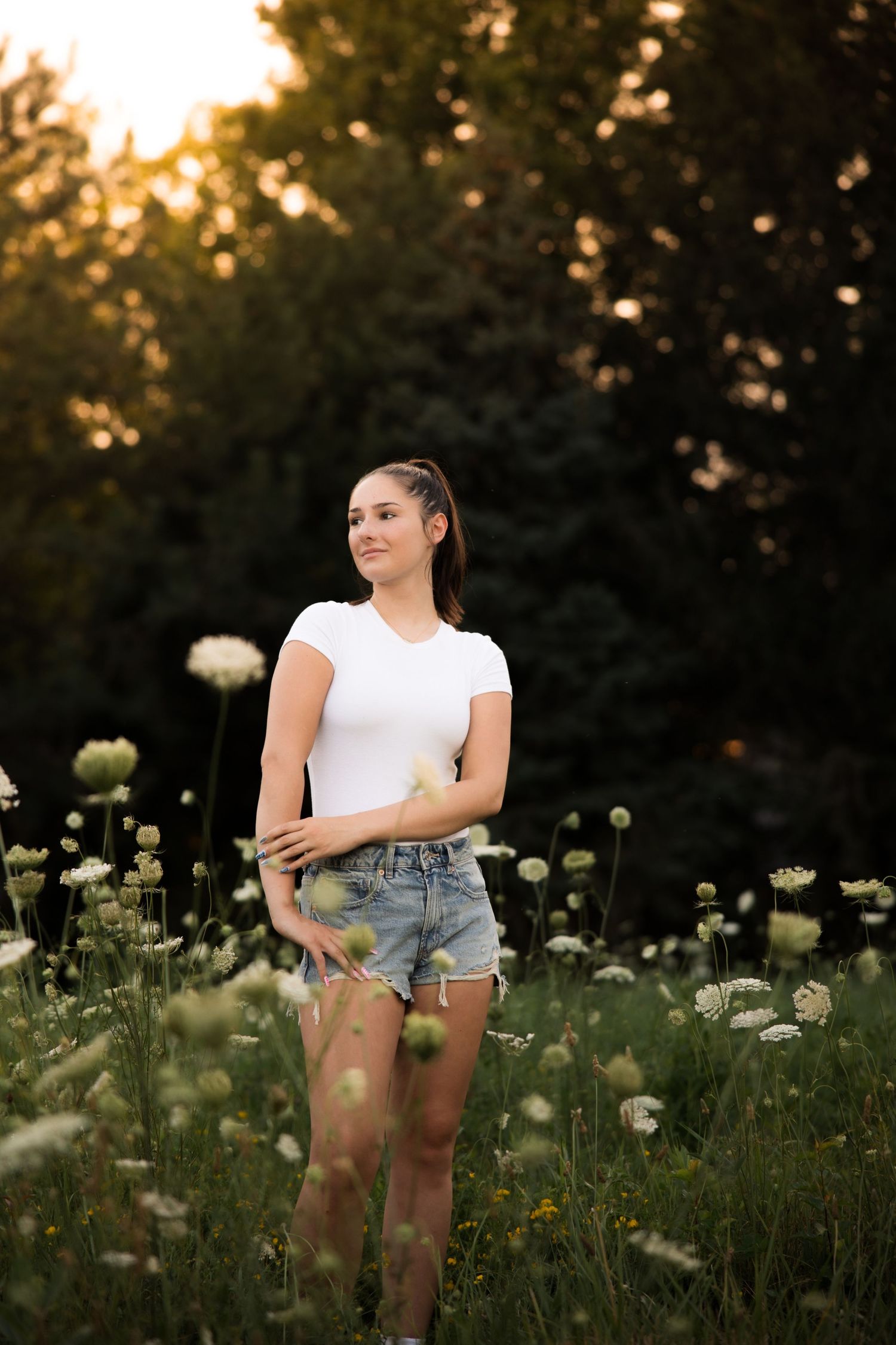 Person in white t-shirt and denim shorts standing in a field of wildflowers at sunset.