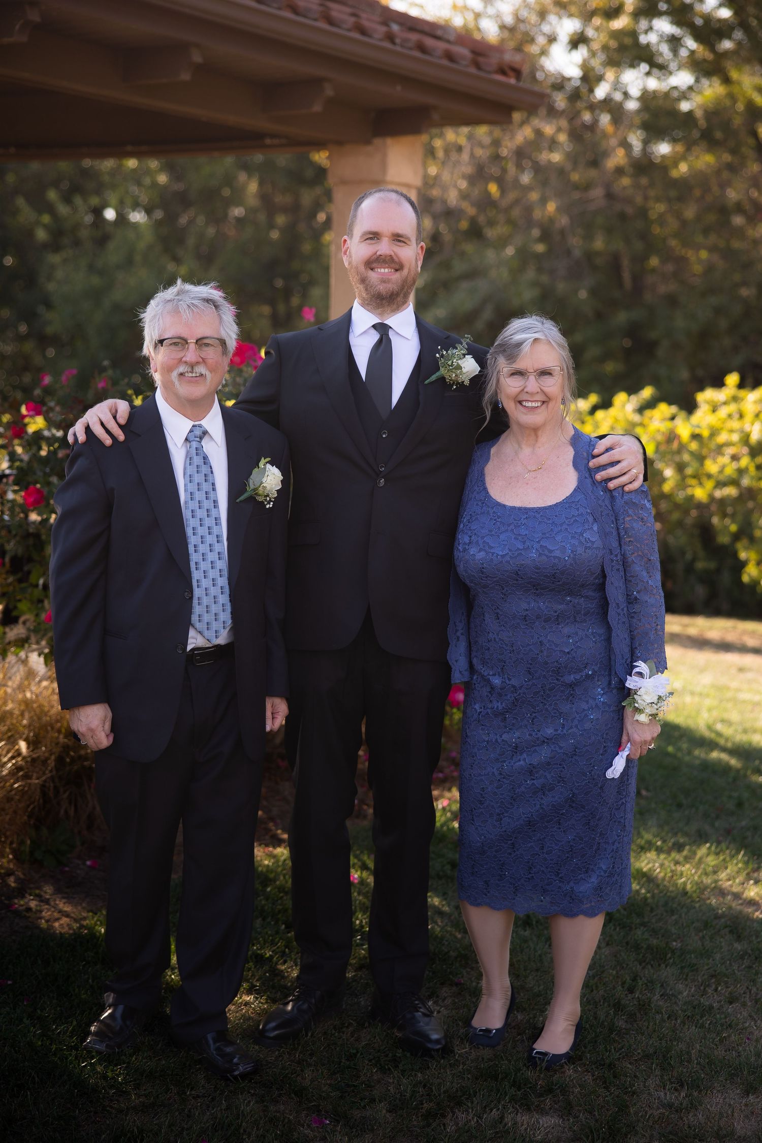 Three people in formal attire stand together outdoors under a gazebo during a special occasion.