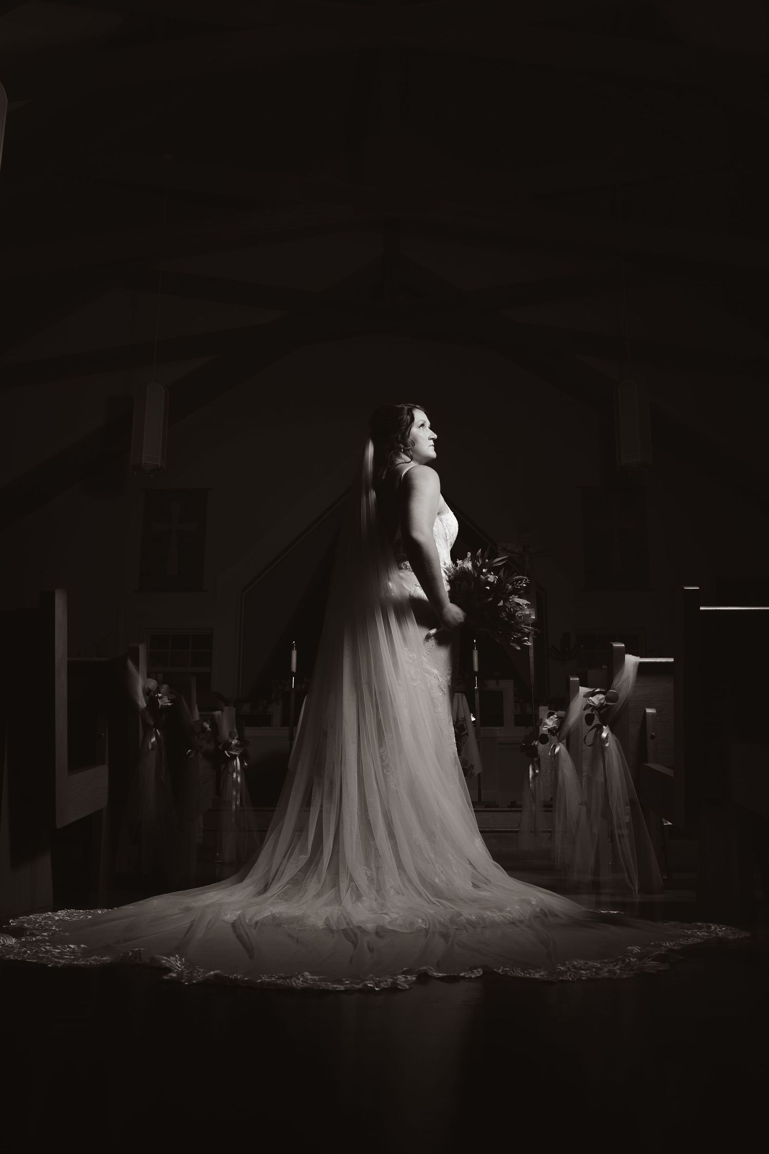 Bride in flowing white gown and veil stands in dramatic black and white church portrait holding bouquet.