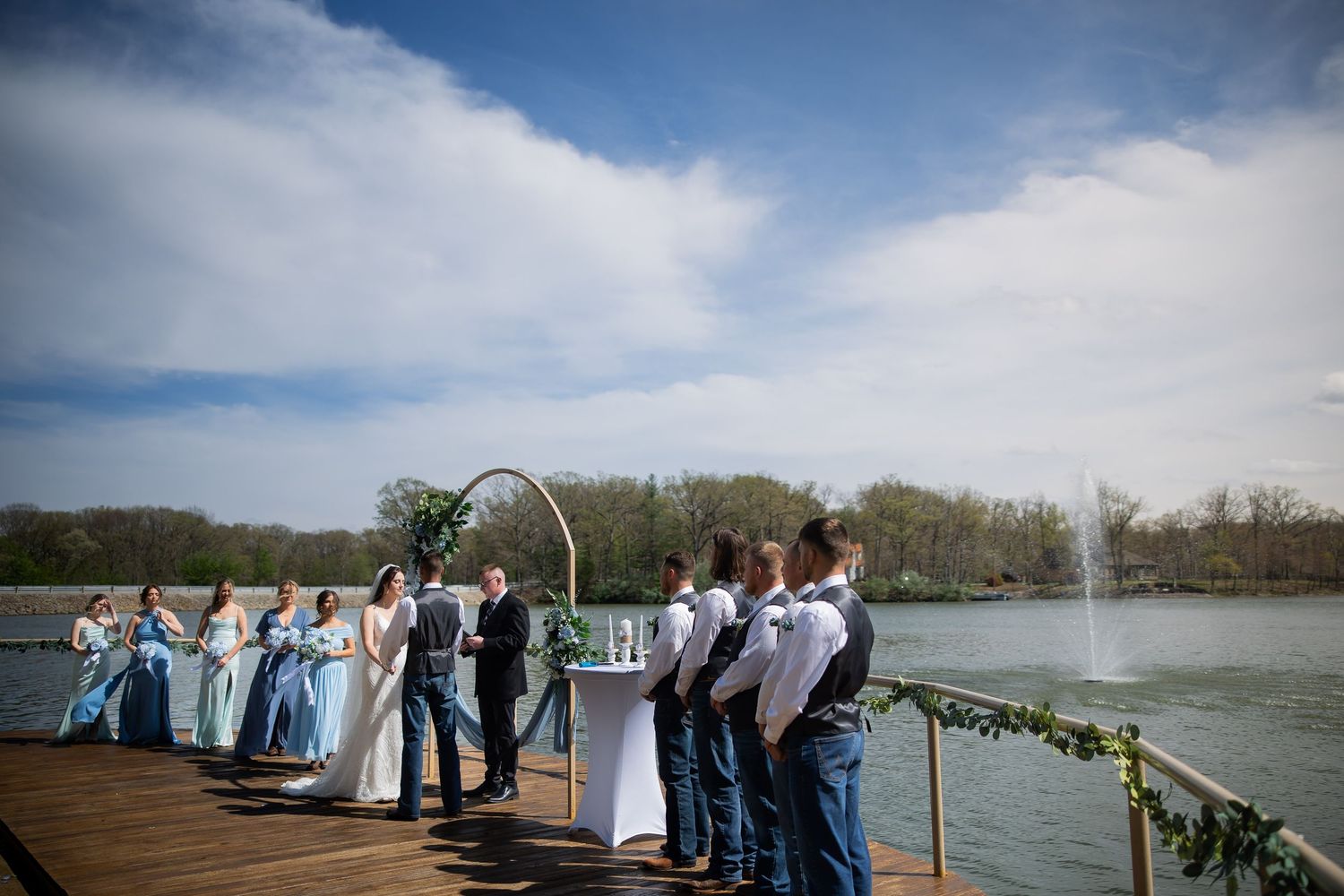 Wedding ceremony on lakeside dock with bridal party, arch, and fountain backdrop under blue sky.