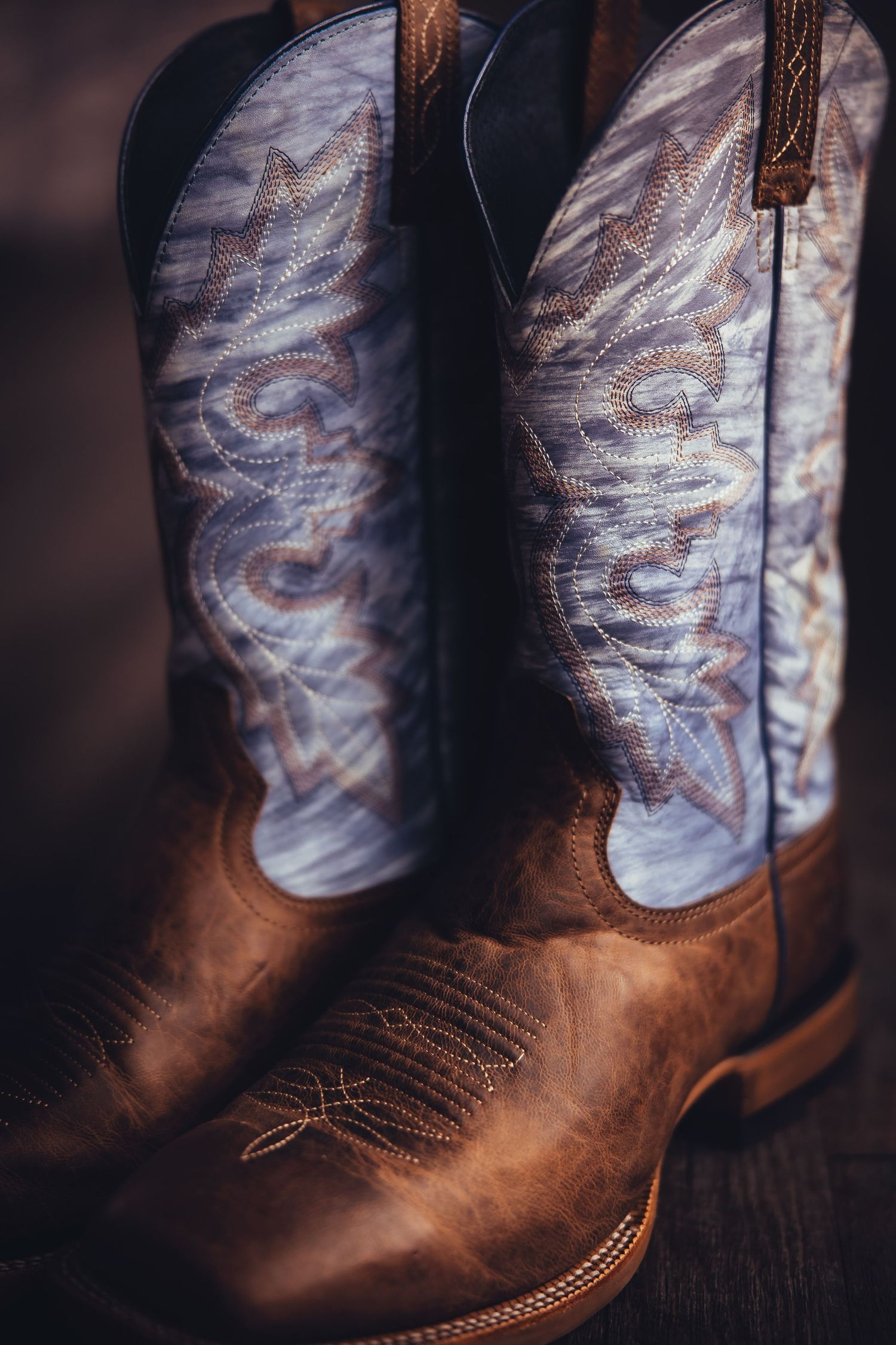 Close-up detail of brown leather cowboy boots with decorative stitching and faded denim blue shaft pattern.
