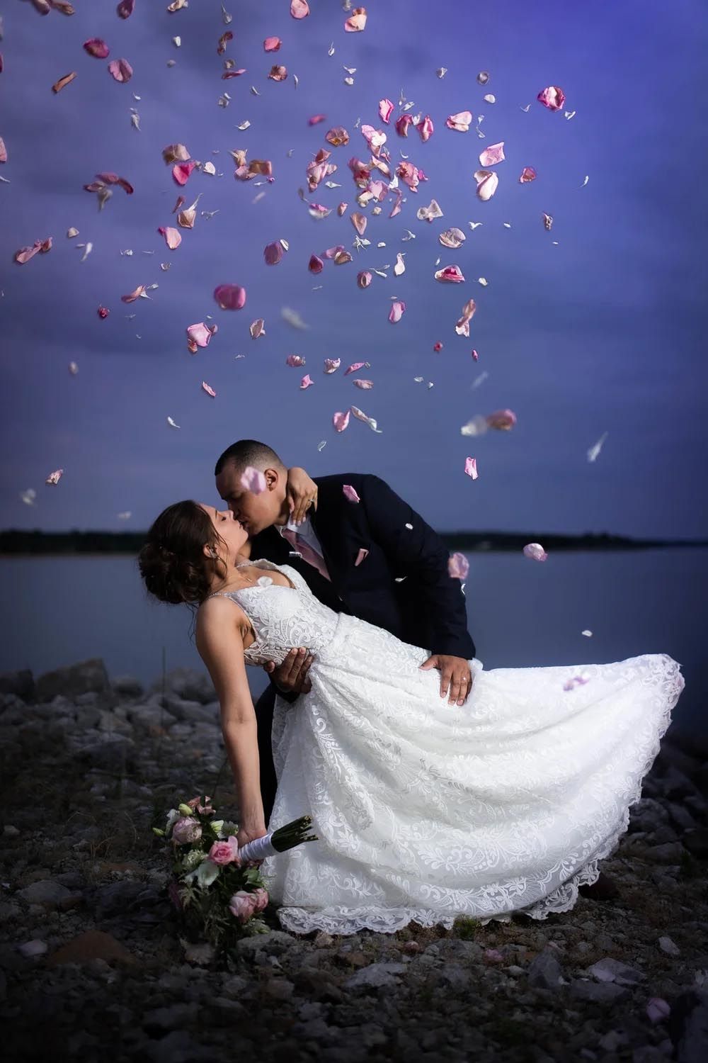 Newlyweds share a romantic dip kiss outdoors at dusk with pink rose petals falling around them, bride in white lace gown.