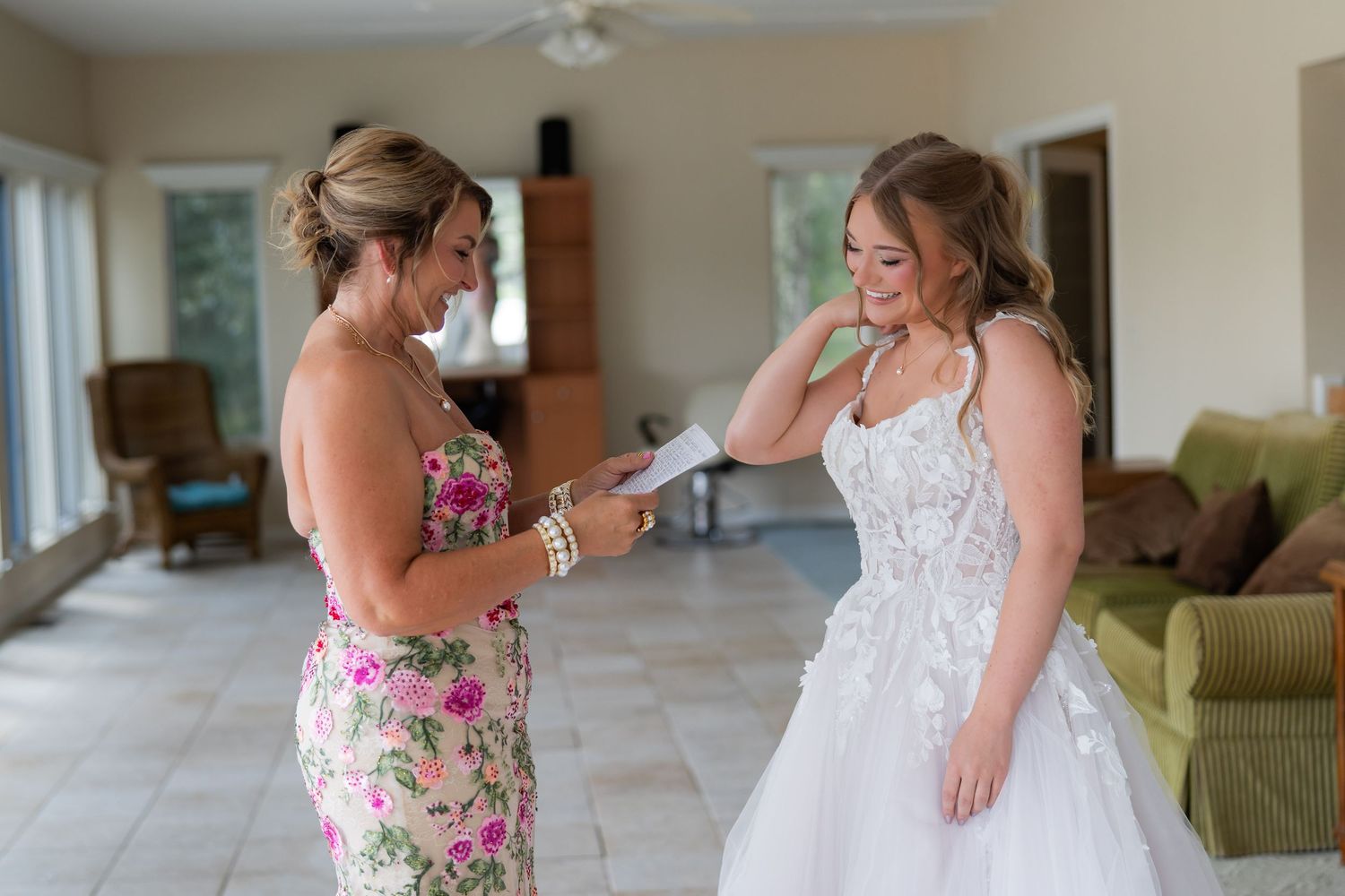 Bride in white lace gown shares emotional moment with woman in floral dress during wedding preparations indoors.
