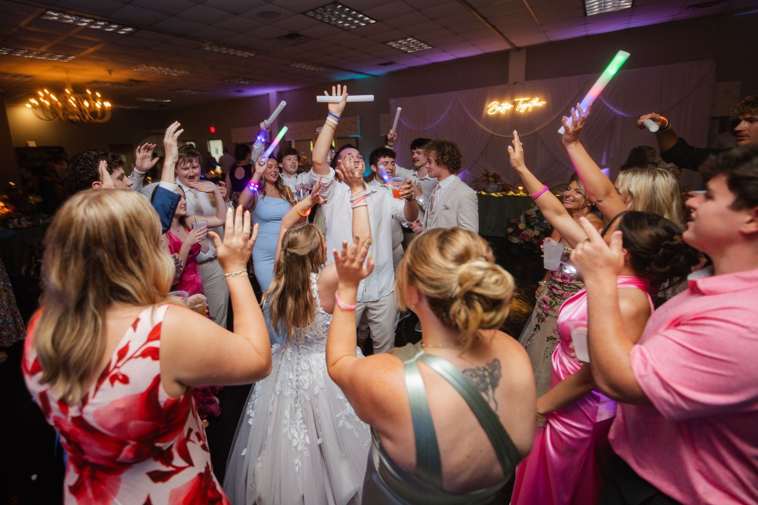 Energetic wedding reception dance floor with guests holding glow sticks under warm lights in a festive ballroom.