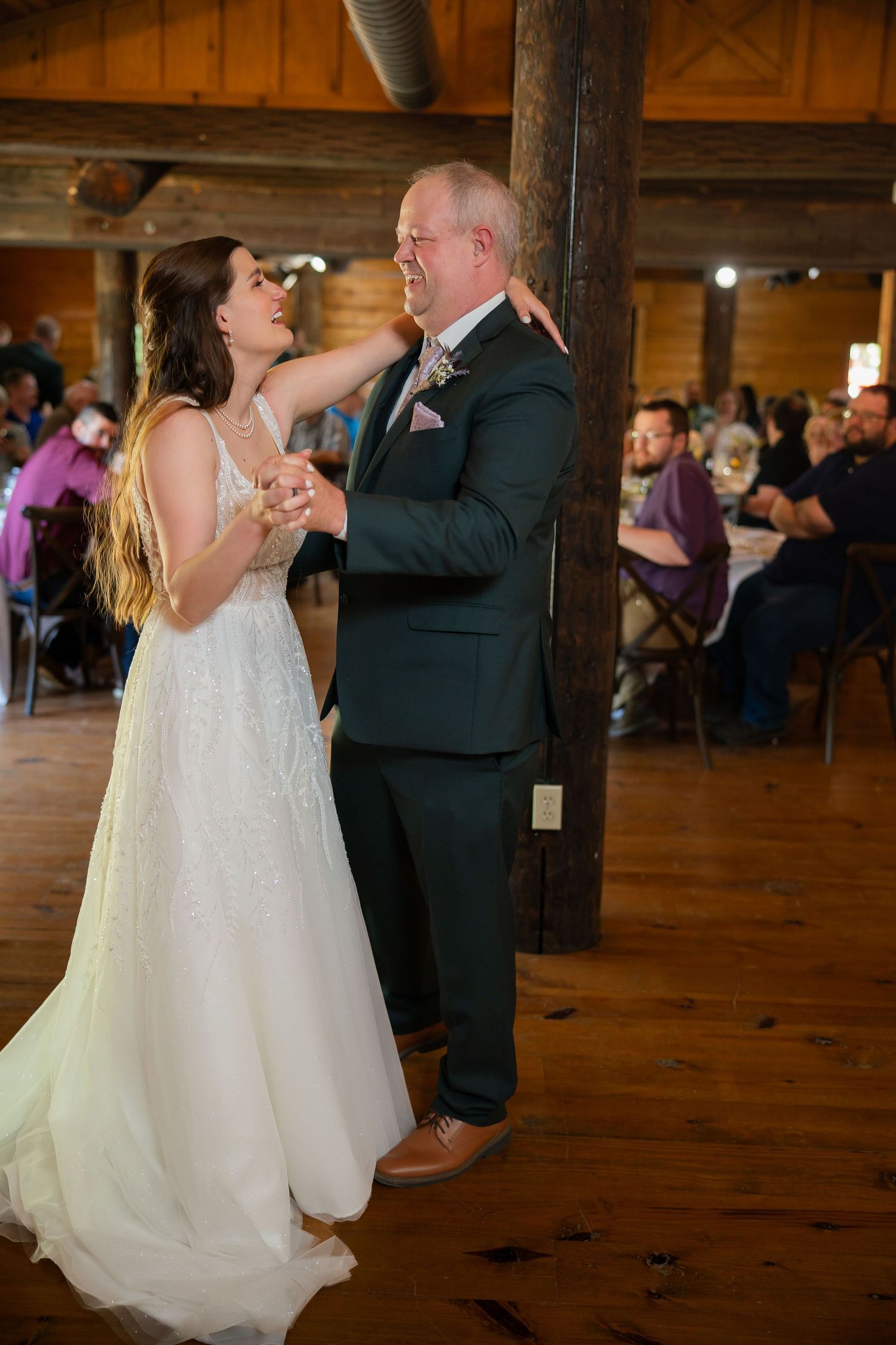 Bride and groom share their first dance in a rustic wooden venue, guests watching in the background.