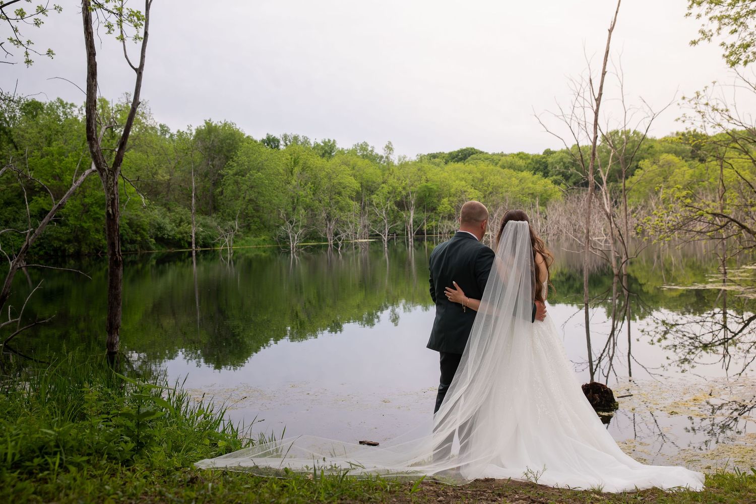 Bride and groom share romantic moment gazing at a peaceful lake surrounded by green trees.