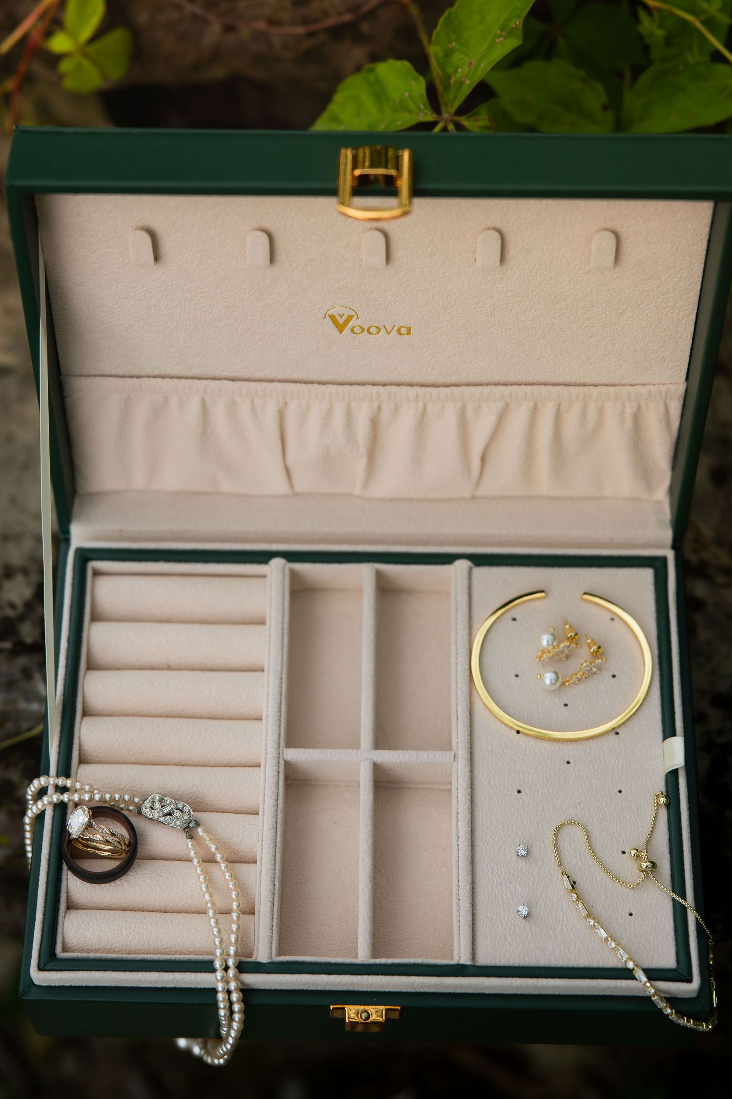 Green Voova jewelry box open displaying gold bangle, pearl earrings, rings, and crystal necklace on cream velvet interior.