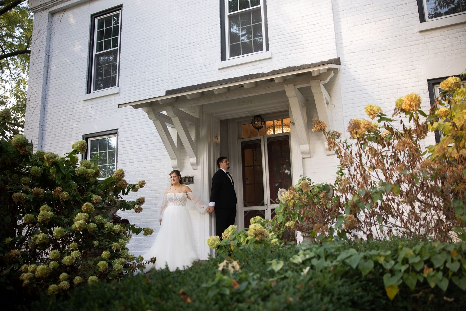 Bride and groom stand at entrance of white colonial wedding venue surrounded by lush greenery and hydrangeas.