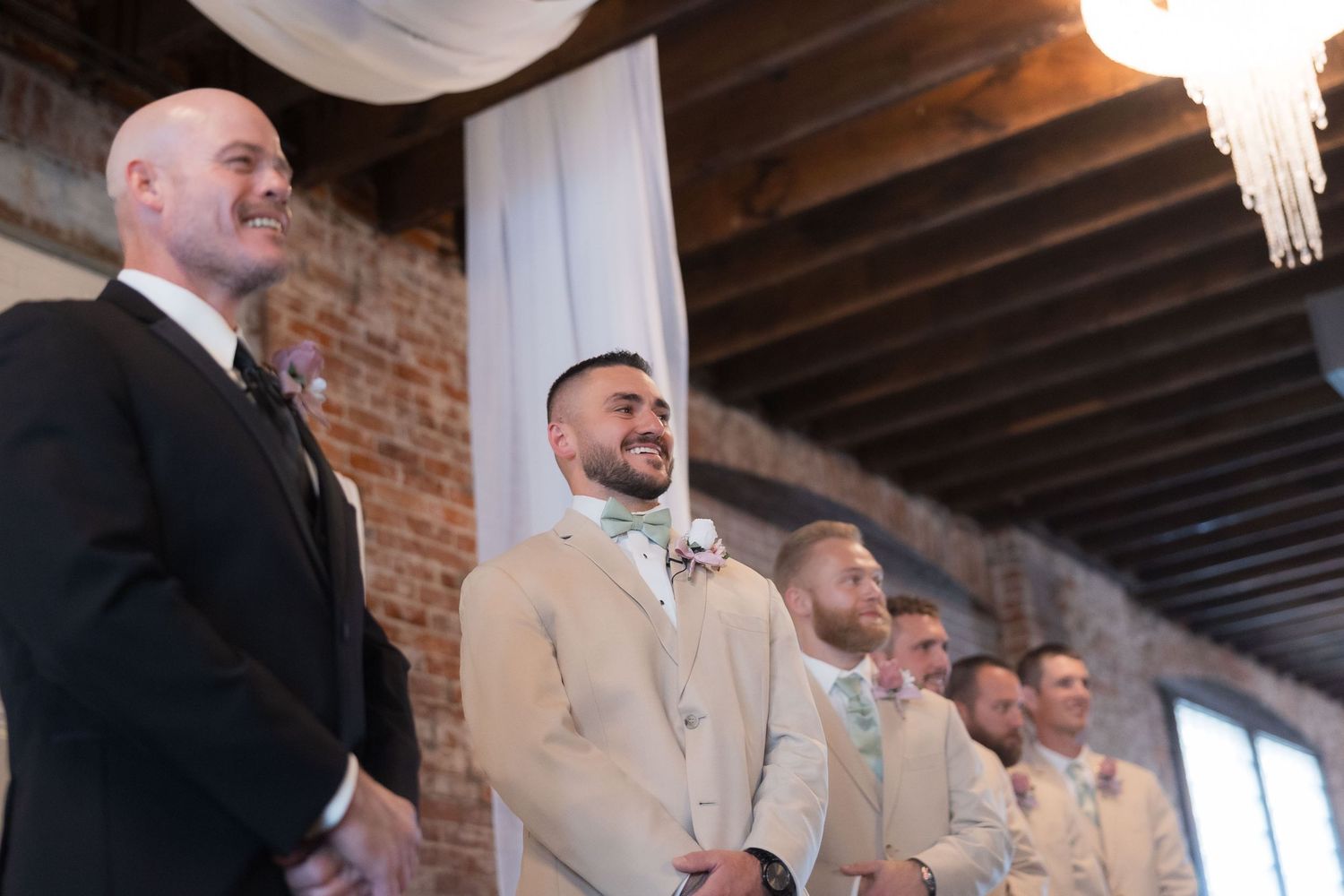 Wedding party stands at altar with groomsmen in light beige suits against exposed brick wall.