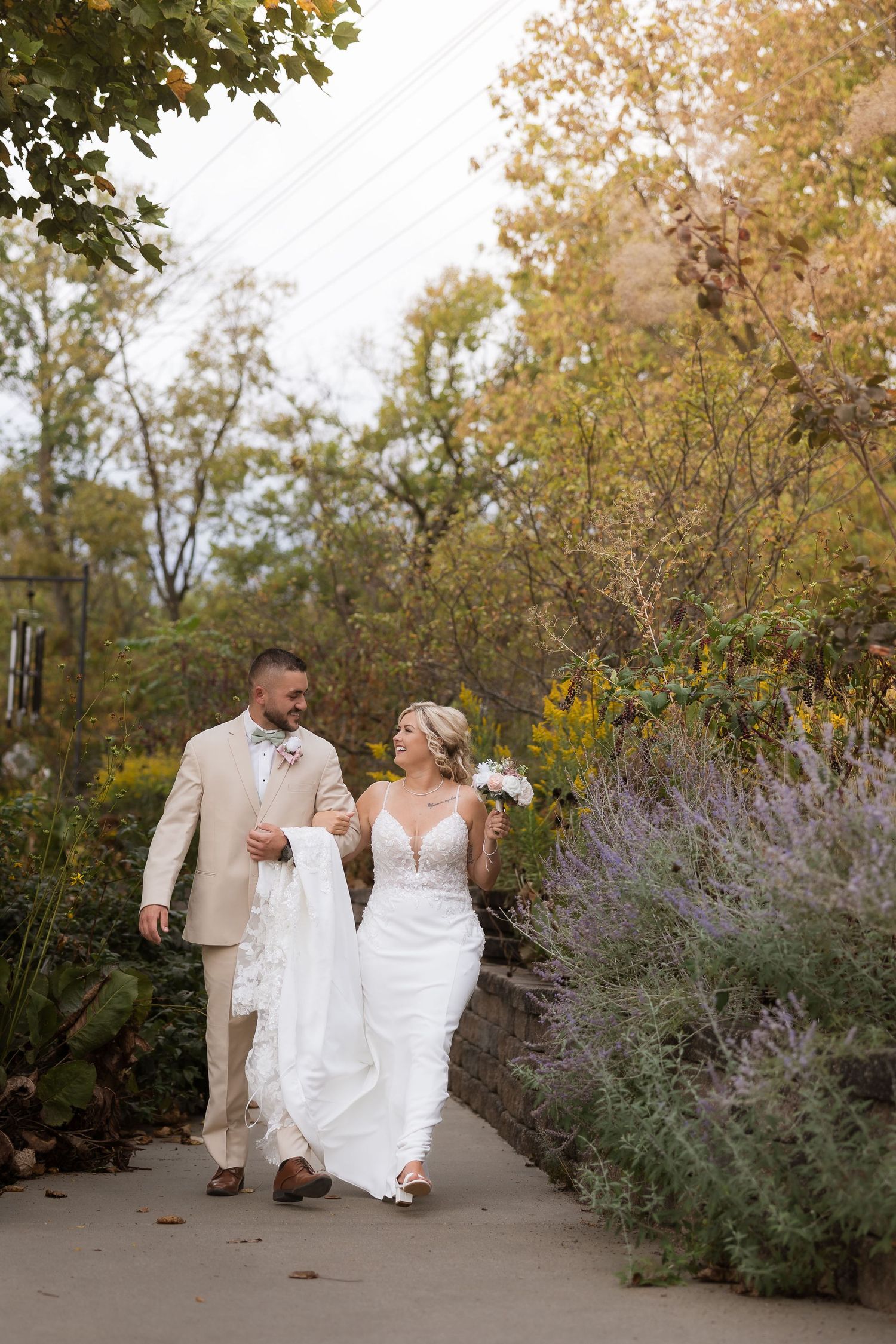 Newlyweds walking through a garden path with autumn foliage and purple flowers.