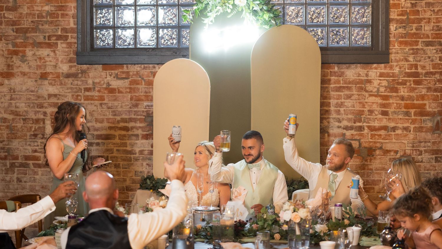 Wedding reception toast in rustic brick venue with arched panels and string lights.