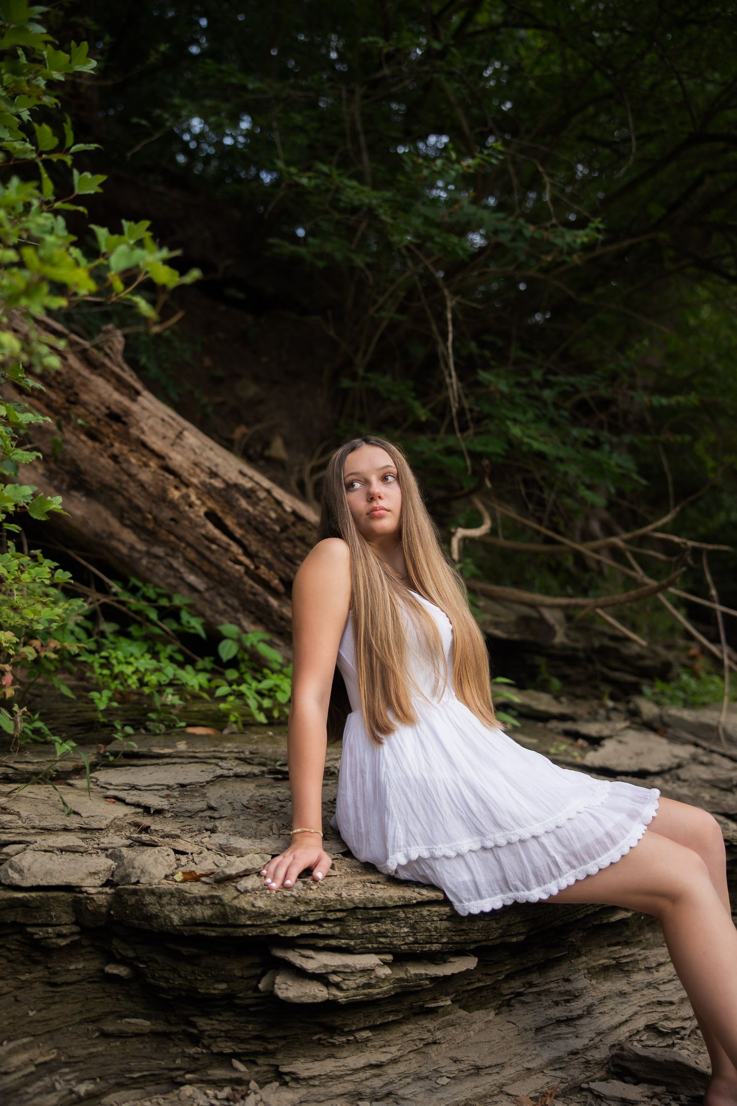 A person in a white dress poses on rocky terrain surrounded by lush greenery in a natural setting.