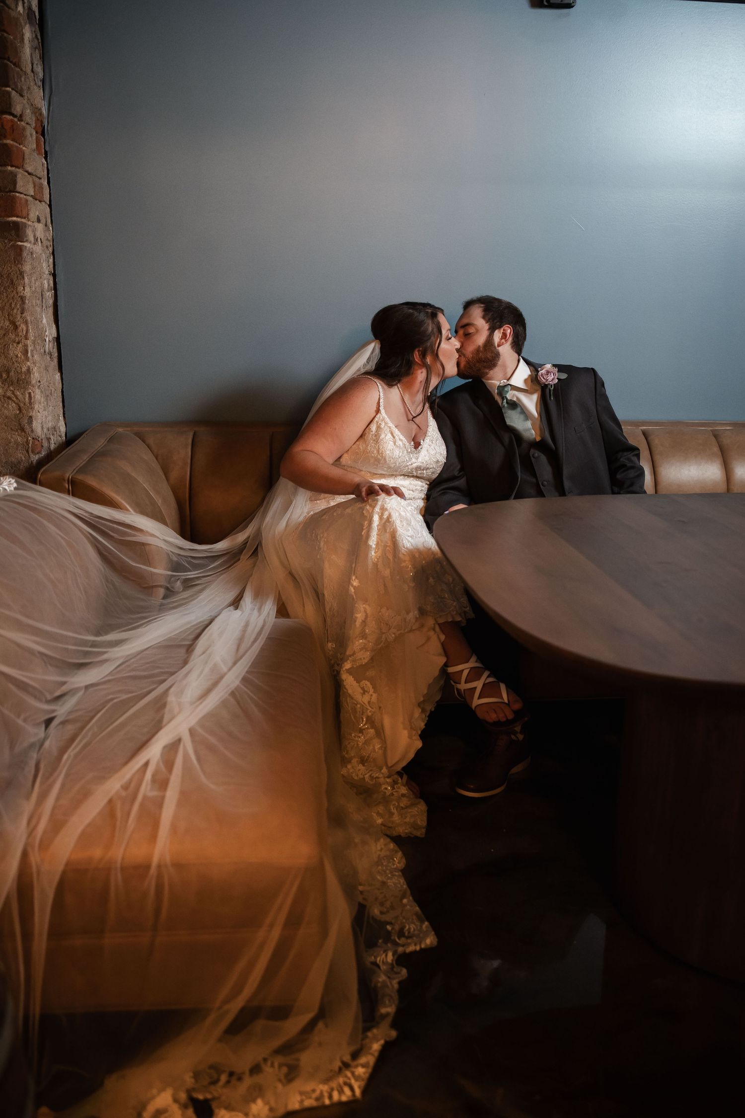 Bride and groom sharing a kiss while seated on a leather sofa, wedding veil flowing gracefully beside them.