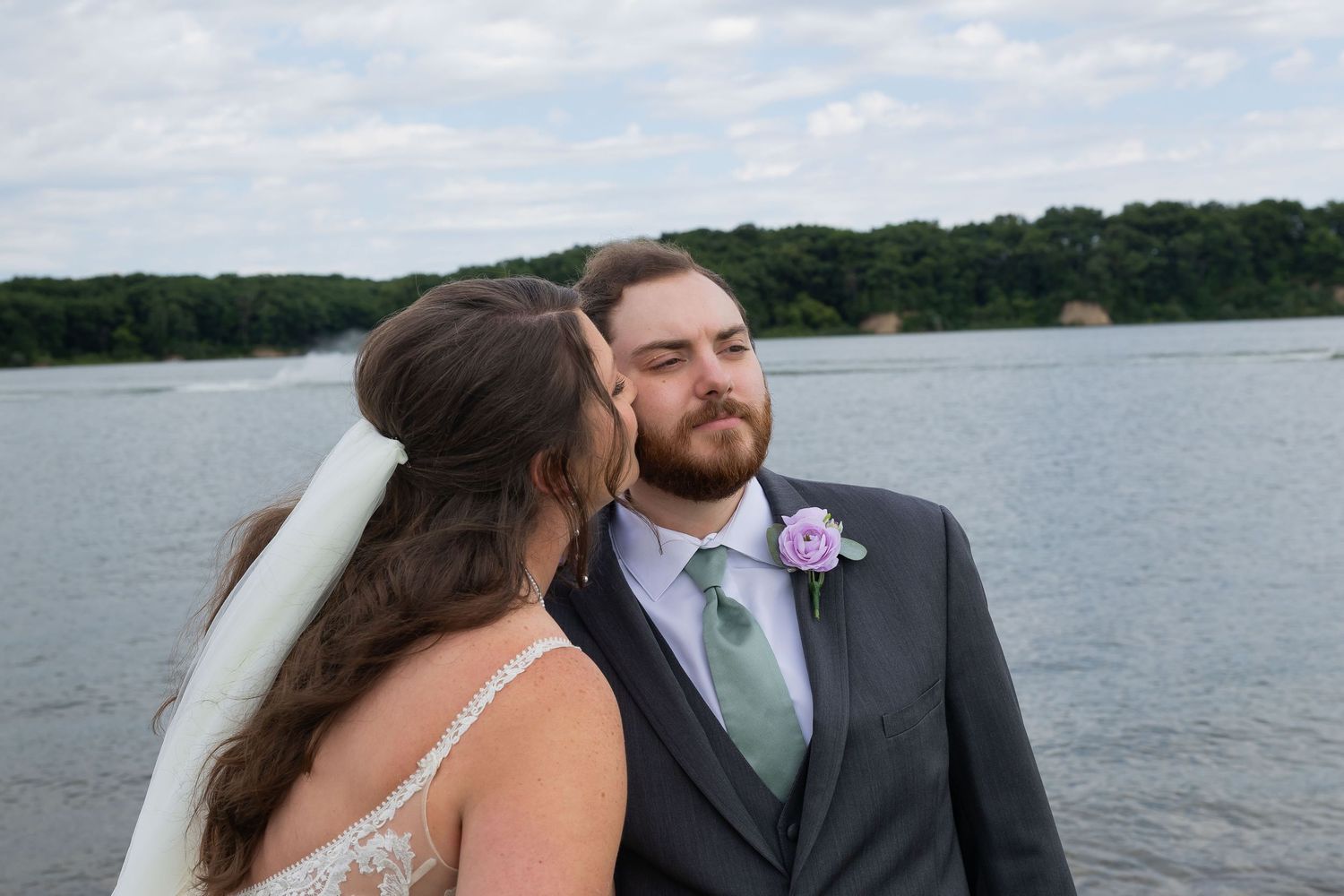 Bride in white veil kissing groom in gray suit with purple boutonniere near a scenic lake backdrop.