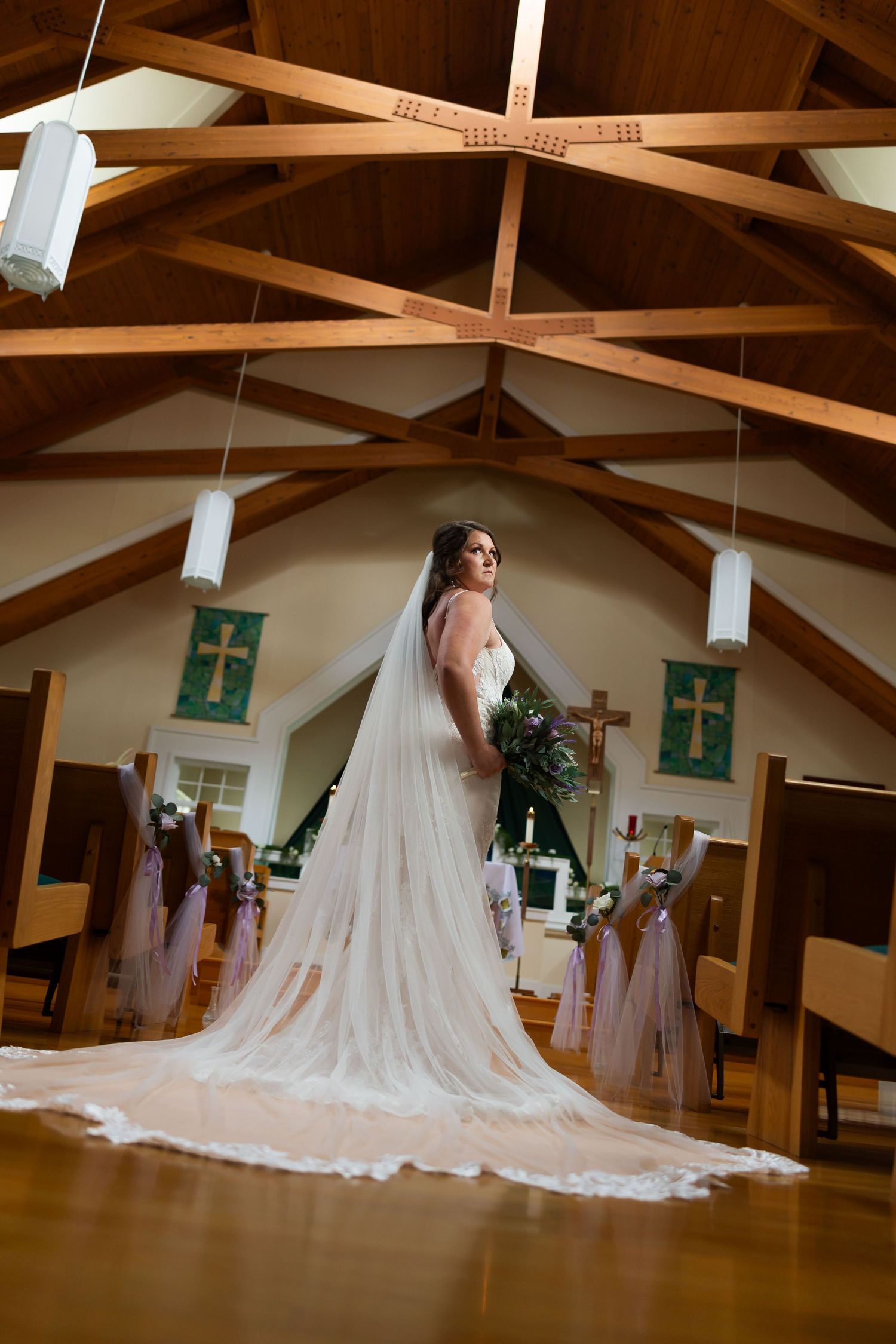 Bride in white gown with long veil stands in church aisle, wooden beamed ceiling and cross decor visible.