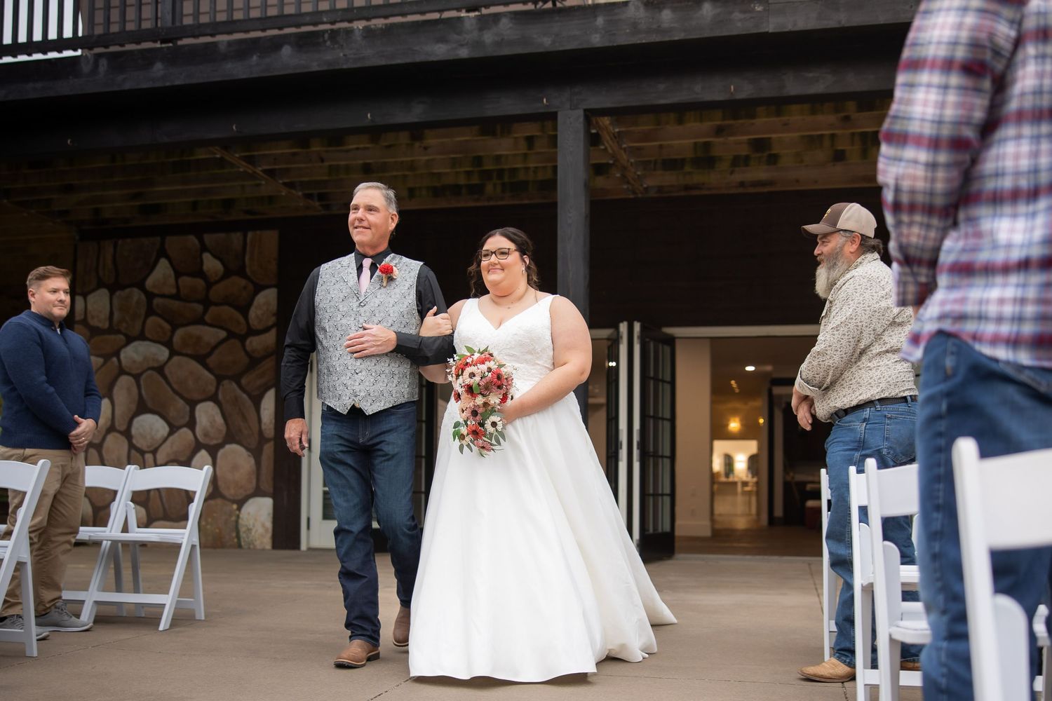 A couple exits their wedding ceremony at a rustic stone and wood venue while guests watch from white folding chairs.