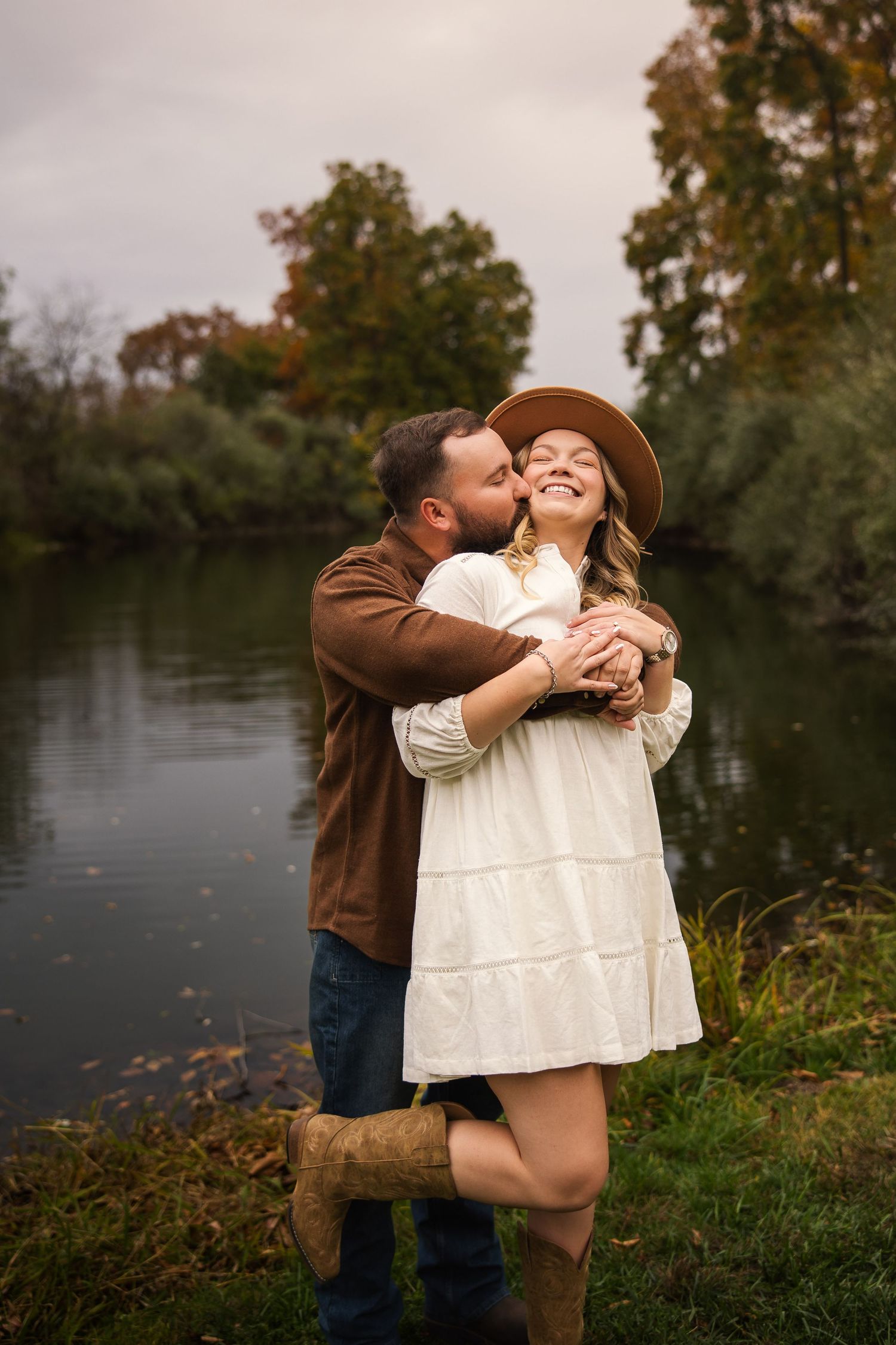 A romantic couple embraces by a tranquil lake during autumn with trees and foliage in the background.