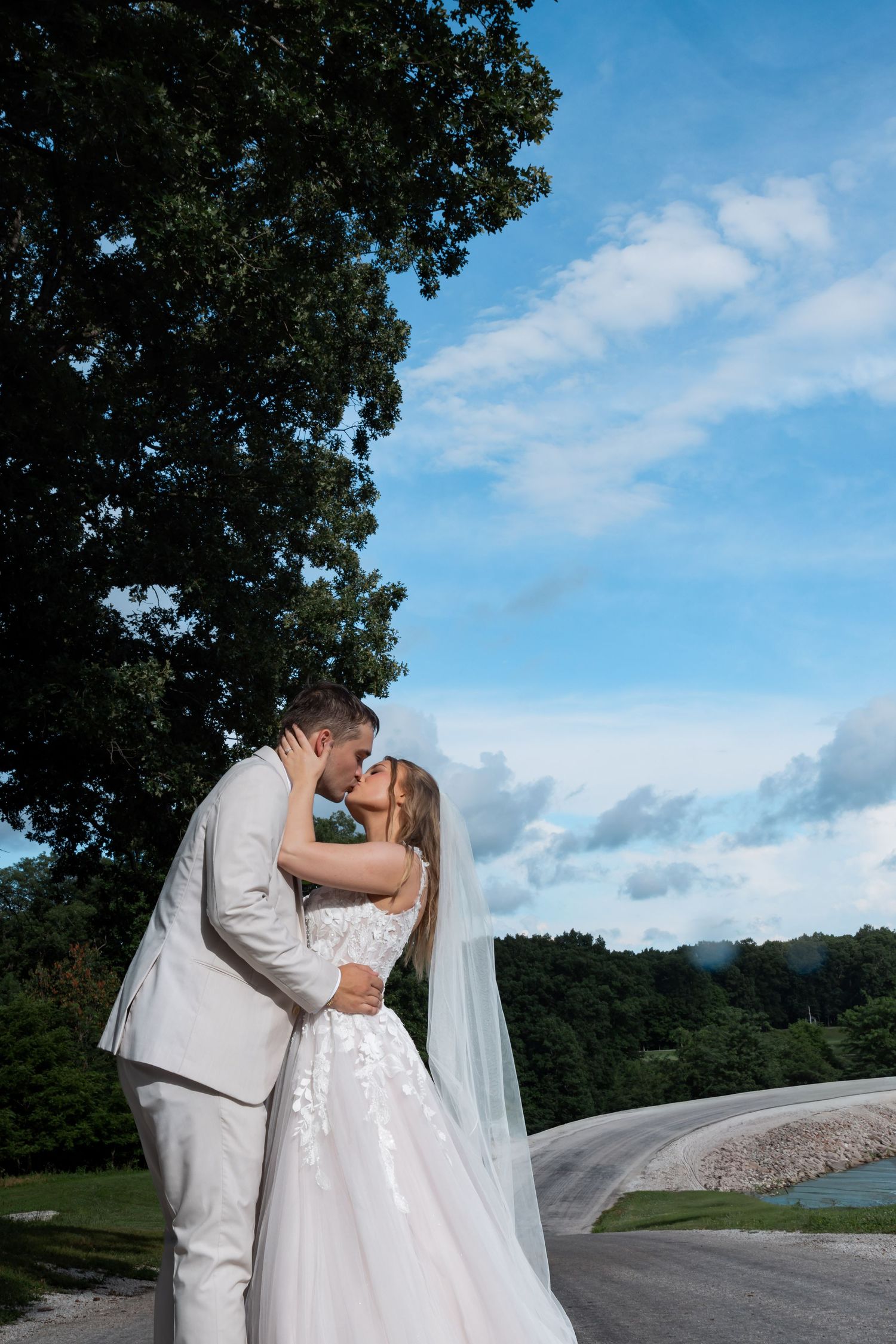 Bride and groom share a romantic kiss outdoors on their wedding day, with a large tree and blue sky behind them.