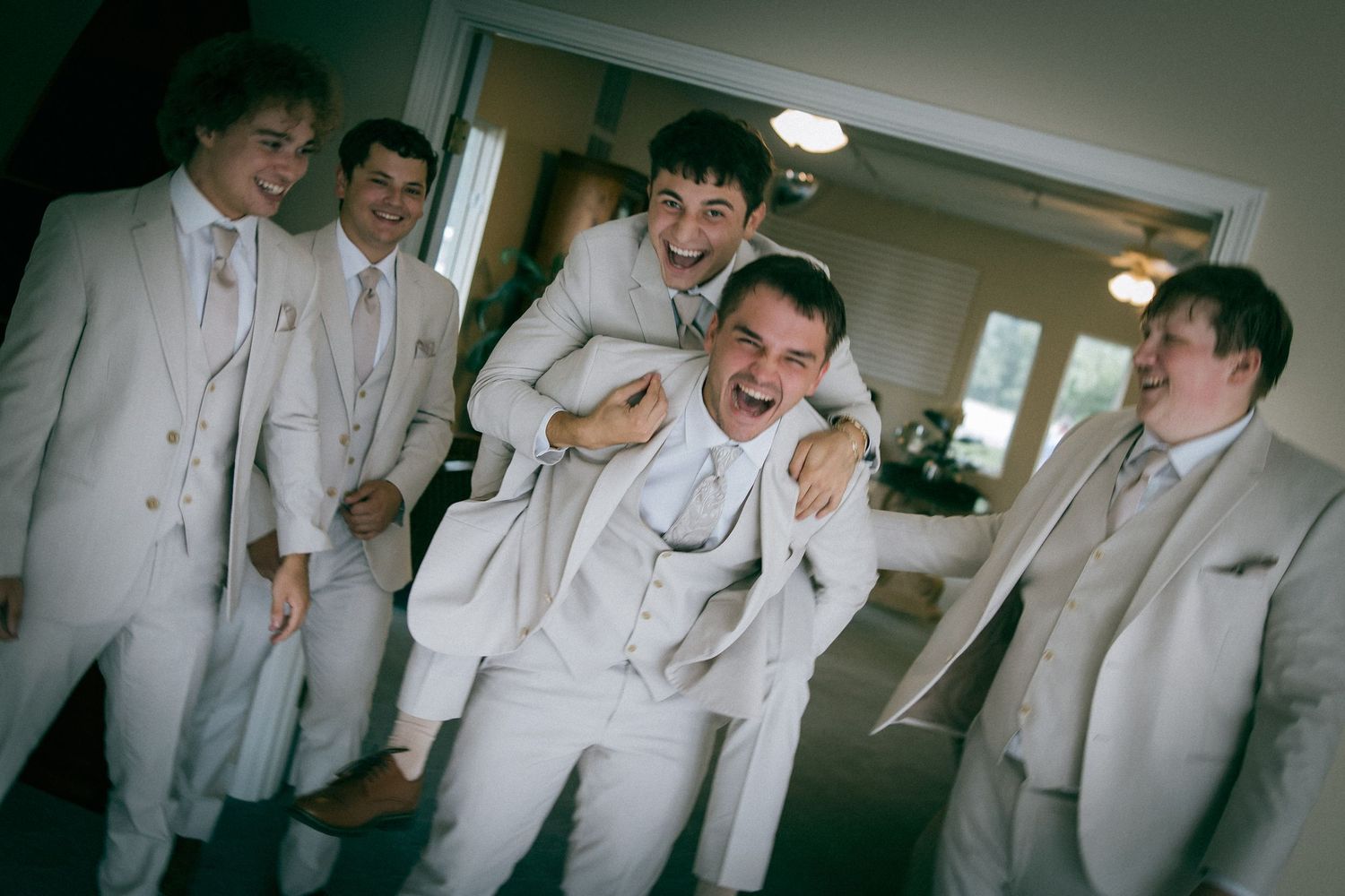 Groomsmen in white suits laughing and joking together before a wedding ceremony indoors.
