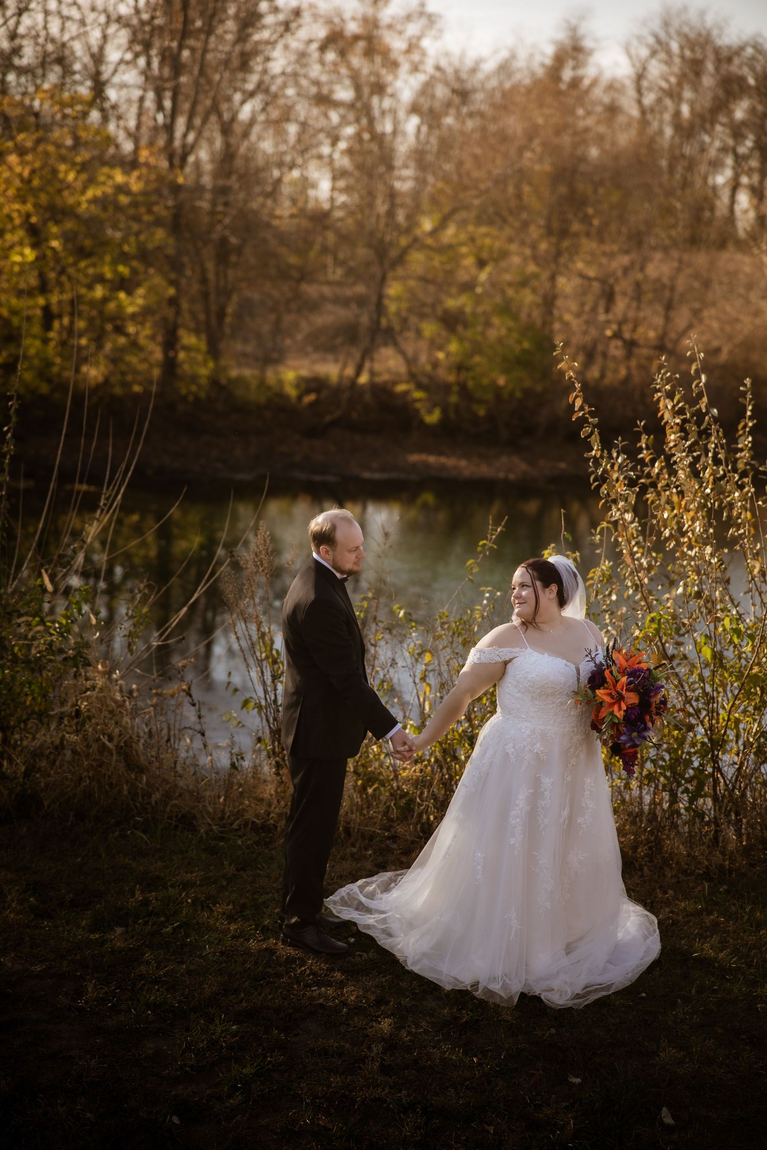 Bride and groom hold hands near a sunlit autumn pond, she in a white lace gown holding a colorful bouquet.