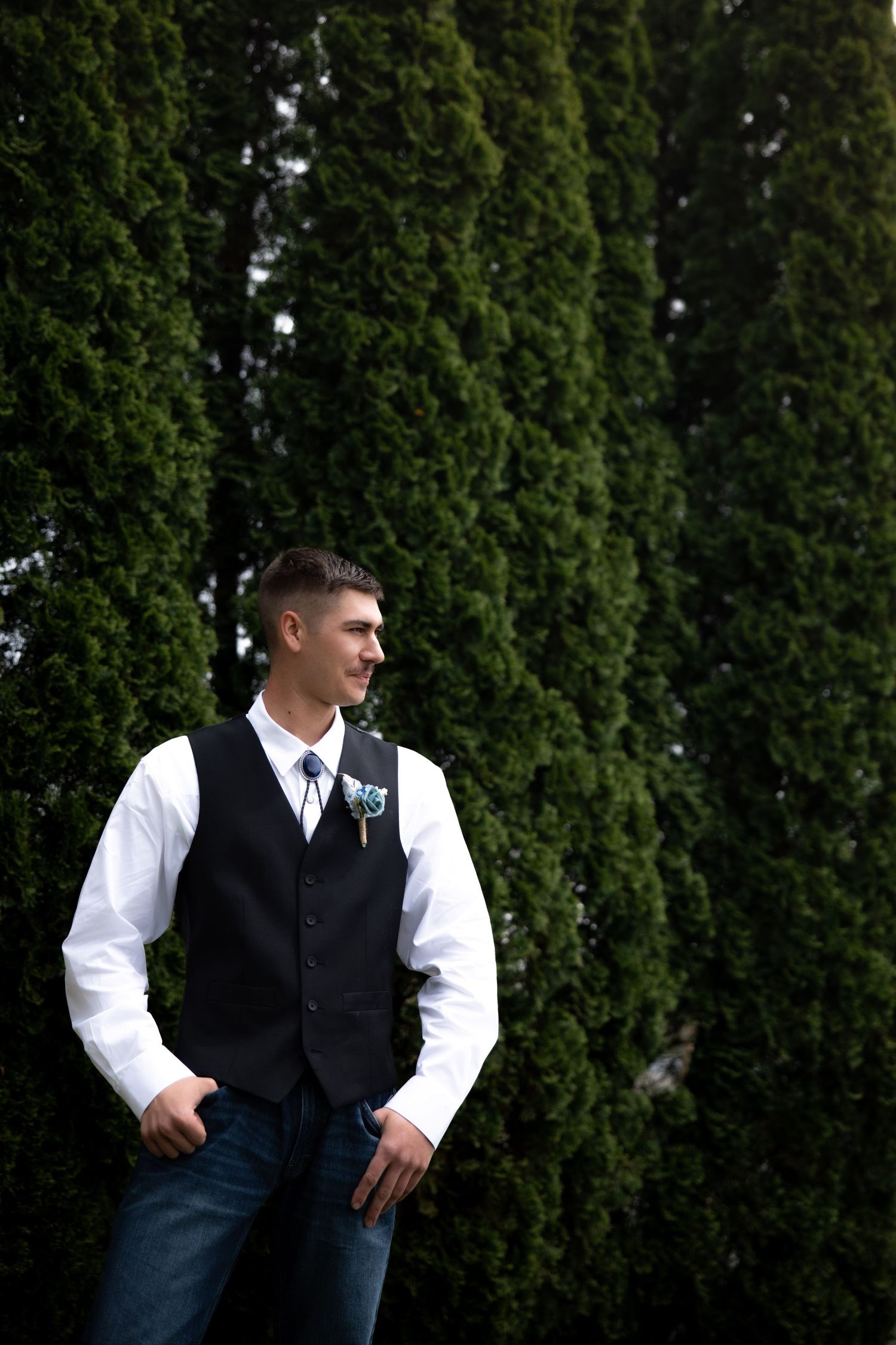 Groom in white shirt, black vest, tie, and jeans stands confidently outdoors before tall green cypress trees.
