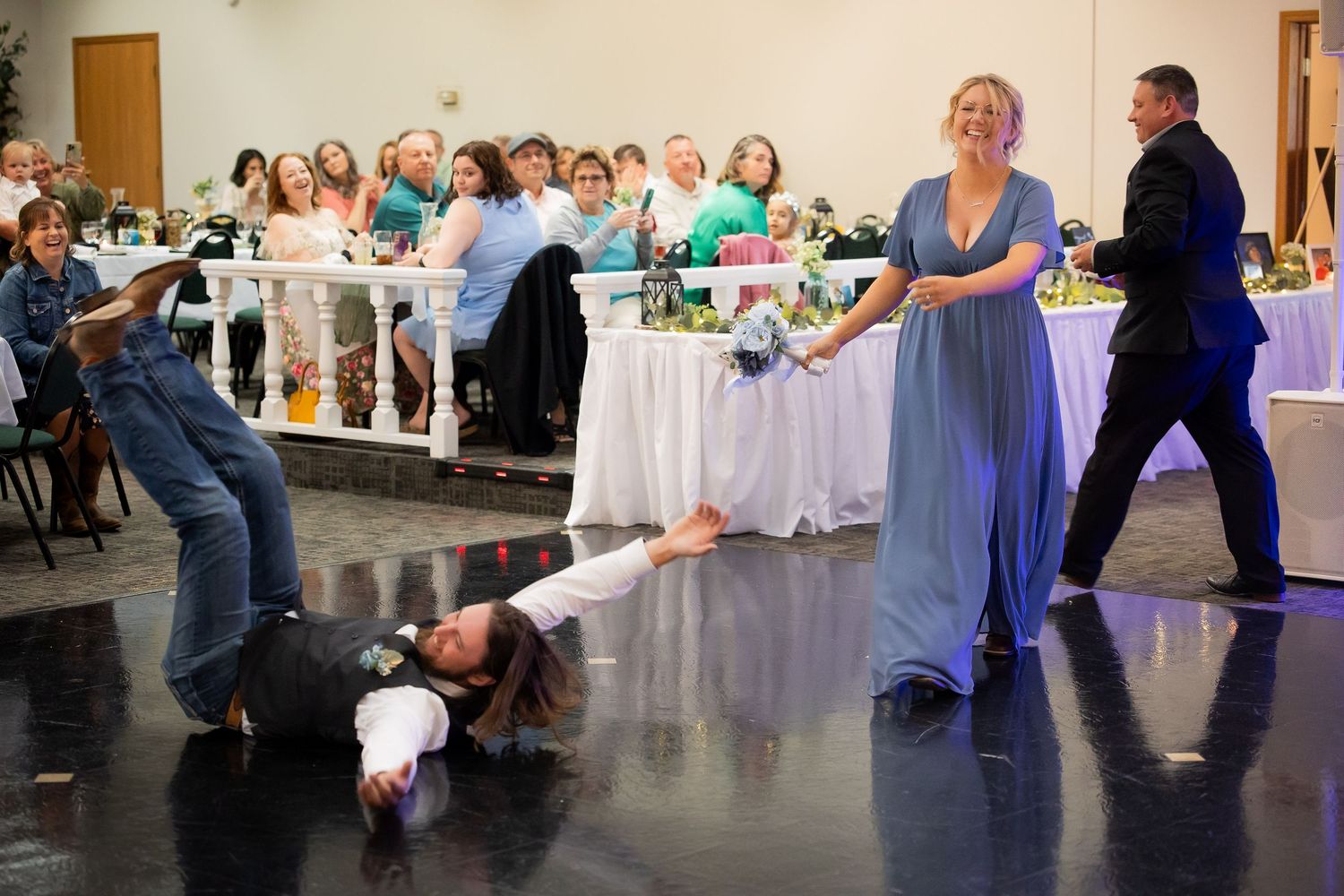 Bridesmaid in blue gown laughs as guest slides across dance floor at lively wedding reception.