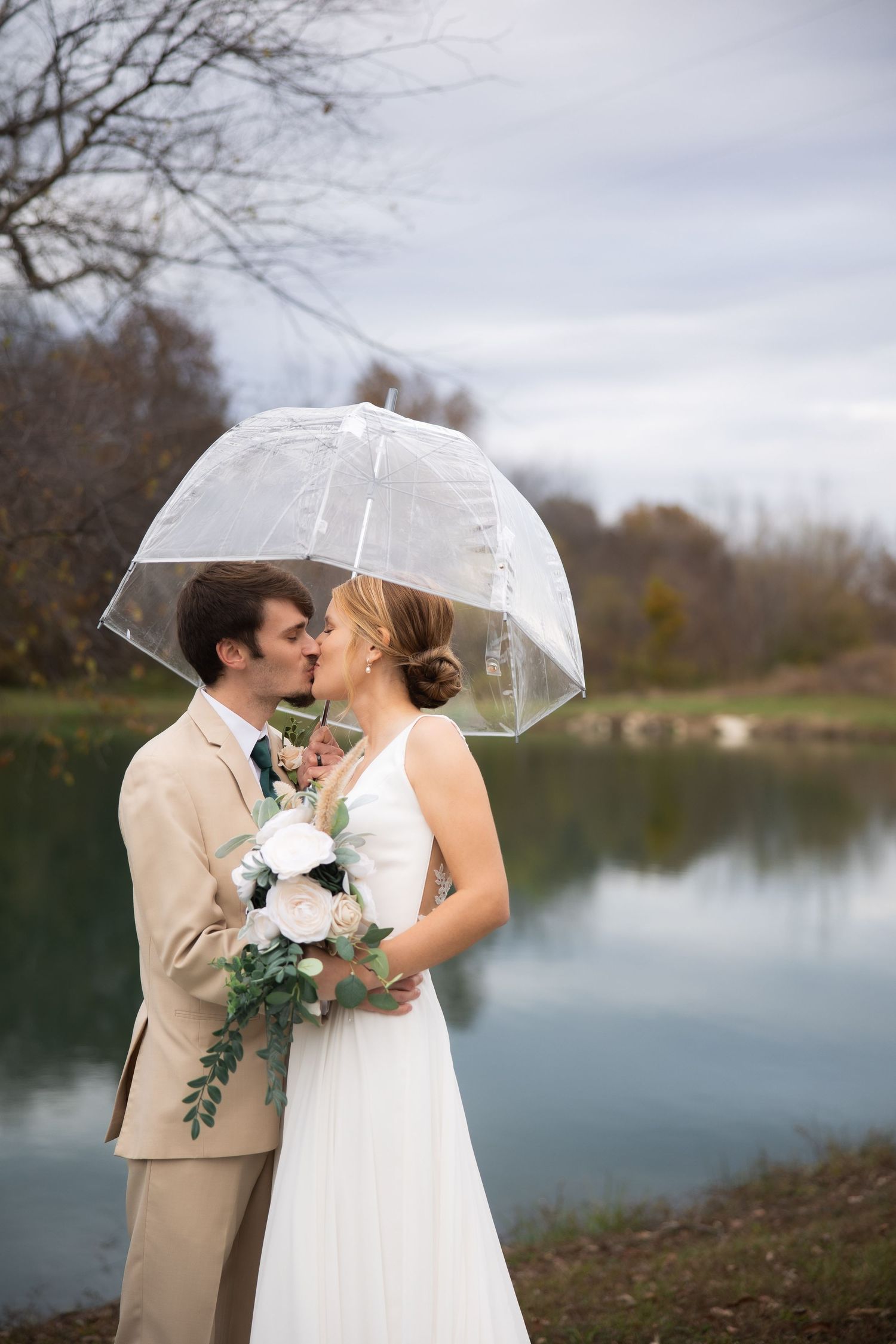 Romantic wedding couple shares intimate moment under clear umbrella by lakeside in autumn.