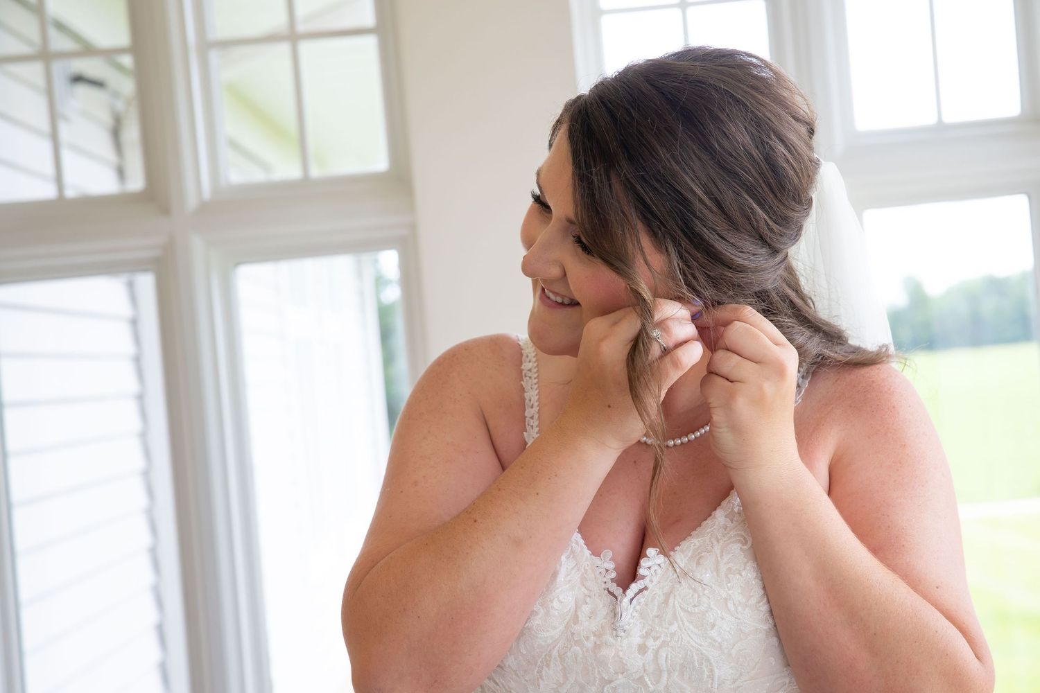 Bride in lace wedding dress putting on pearl earrings while getting ready near bright windows.