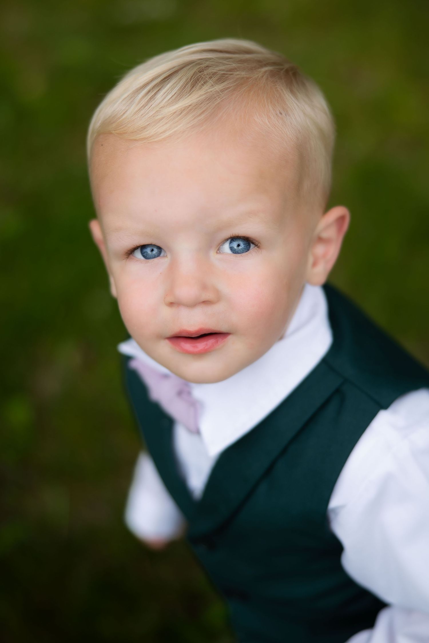 Adorable blonde toddler boy with blue eyes wearing dark green vest and white dress shirt at outdoor event.