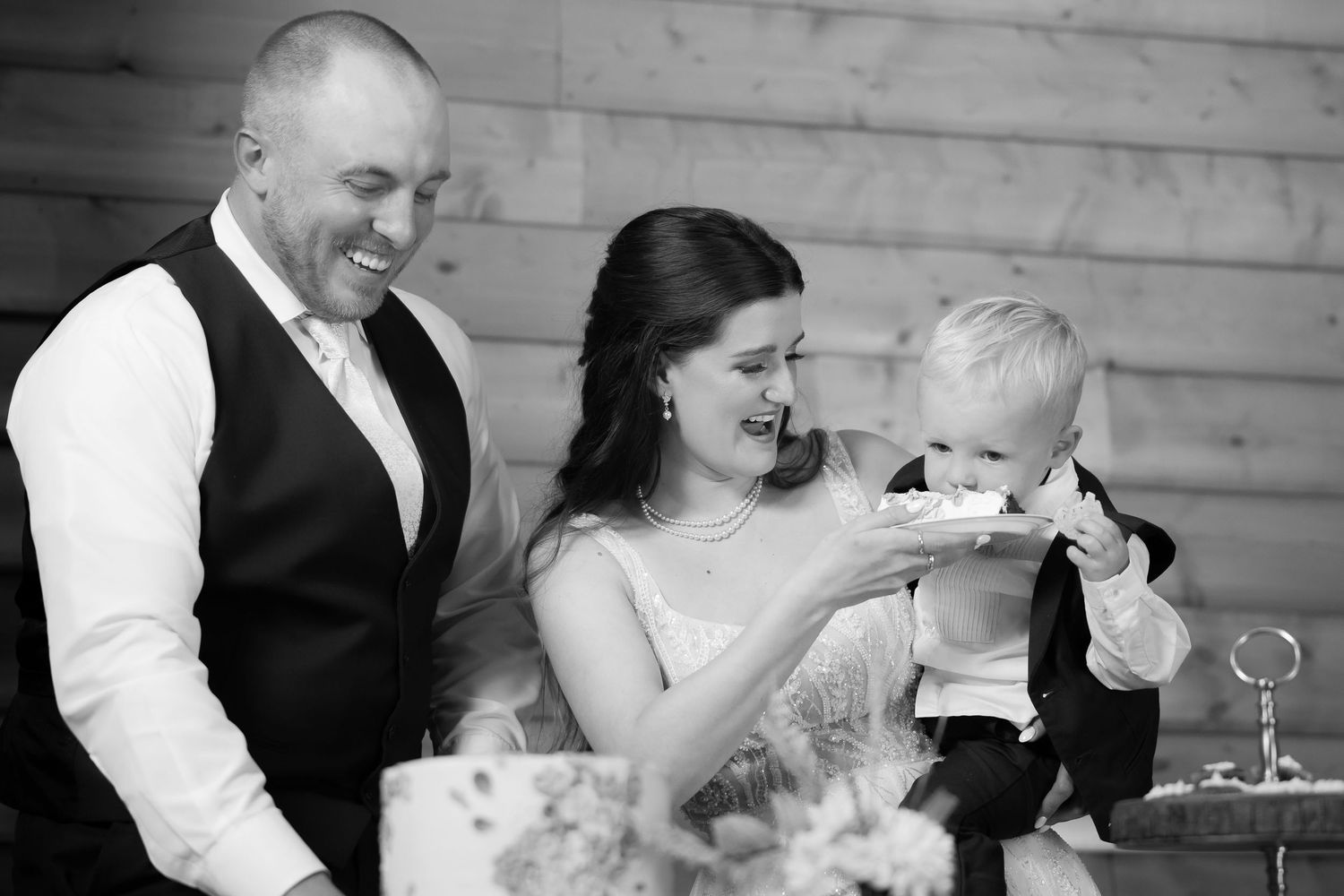 Black and white photo of bride and groom smiling as they feed wedding cake to a baby.