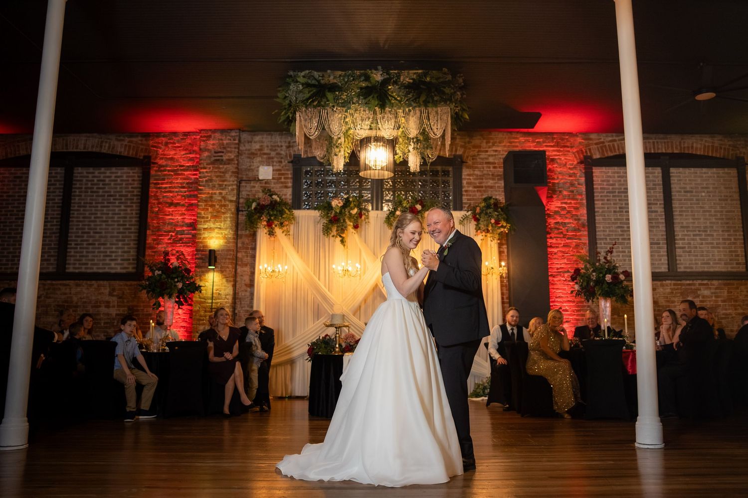 Bride and groom share first dance on elegant ballroom floor with red uplighting and floral chandelier backdrop.