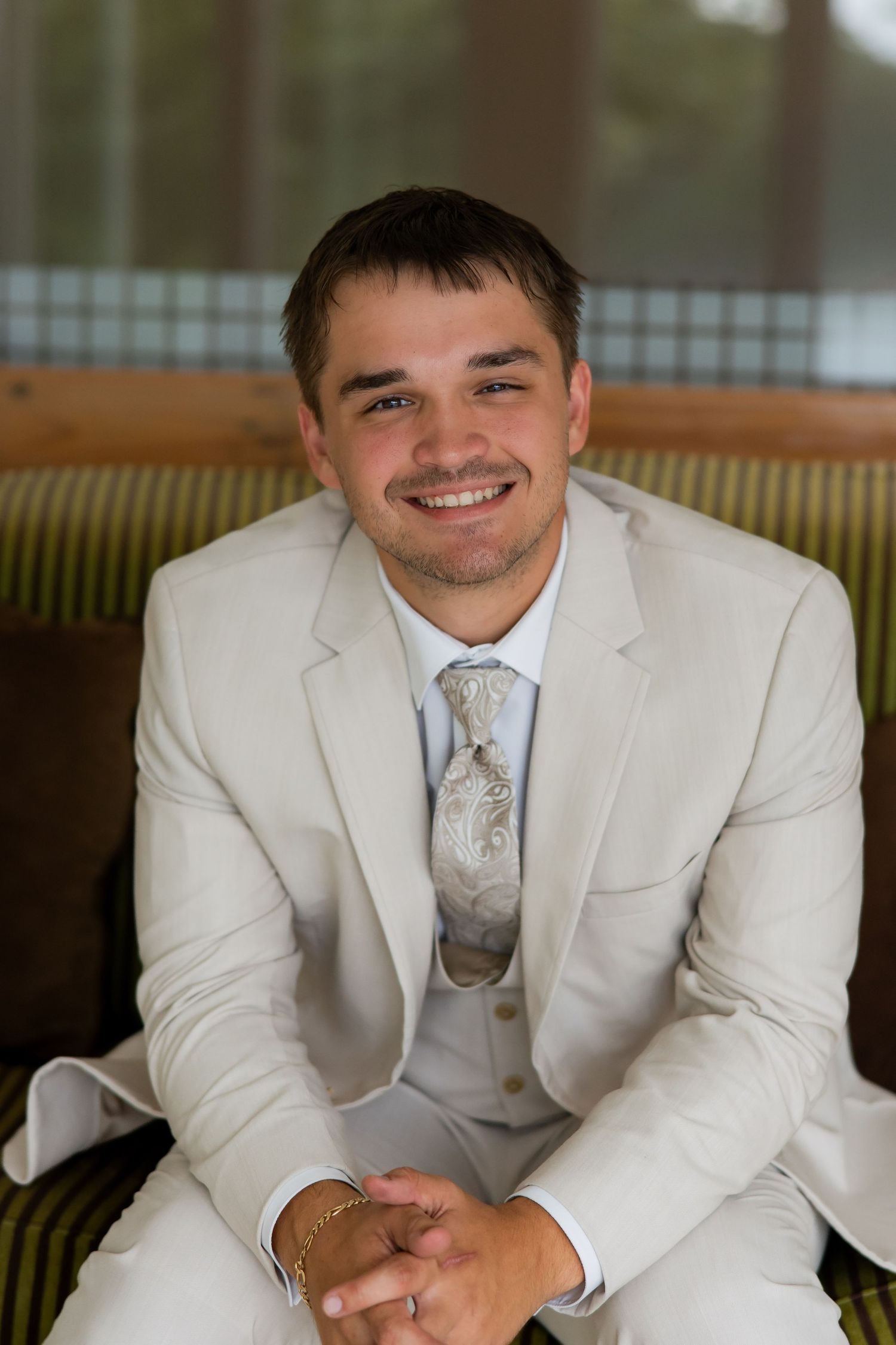 Smiling groom in a cream suit and paisley tie sitting on a striped chair at a wedding venue.