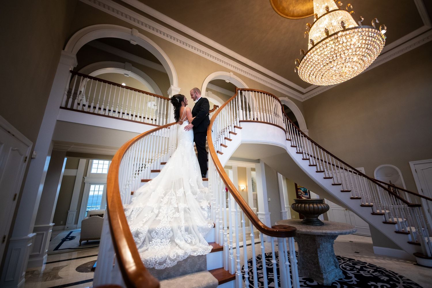 Bride and groom share a romantic moment on grand double staircase beneath crystal chandelier in elegant wedding venue.