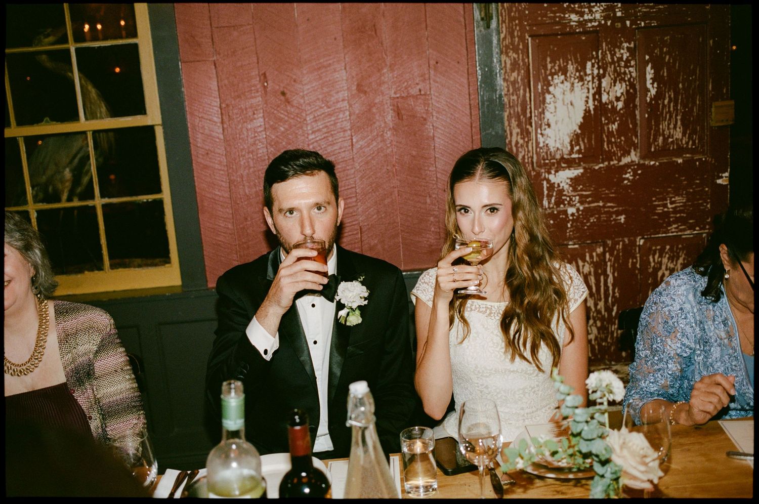 Bride and groom sipping drinks at wedding reception table, rustic red barn wood wall backdrop, candid film photography.