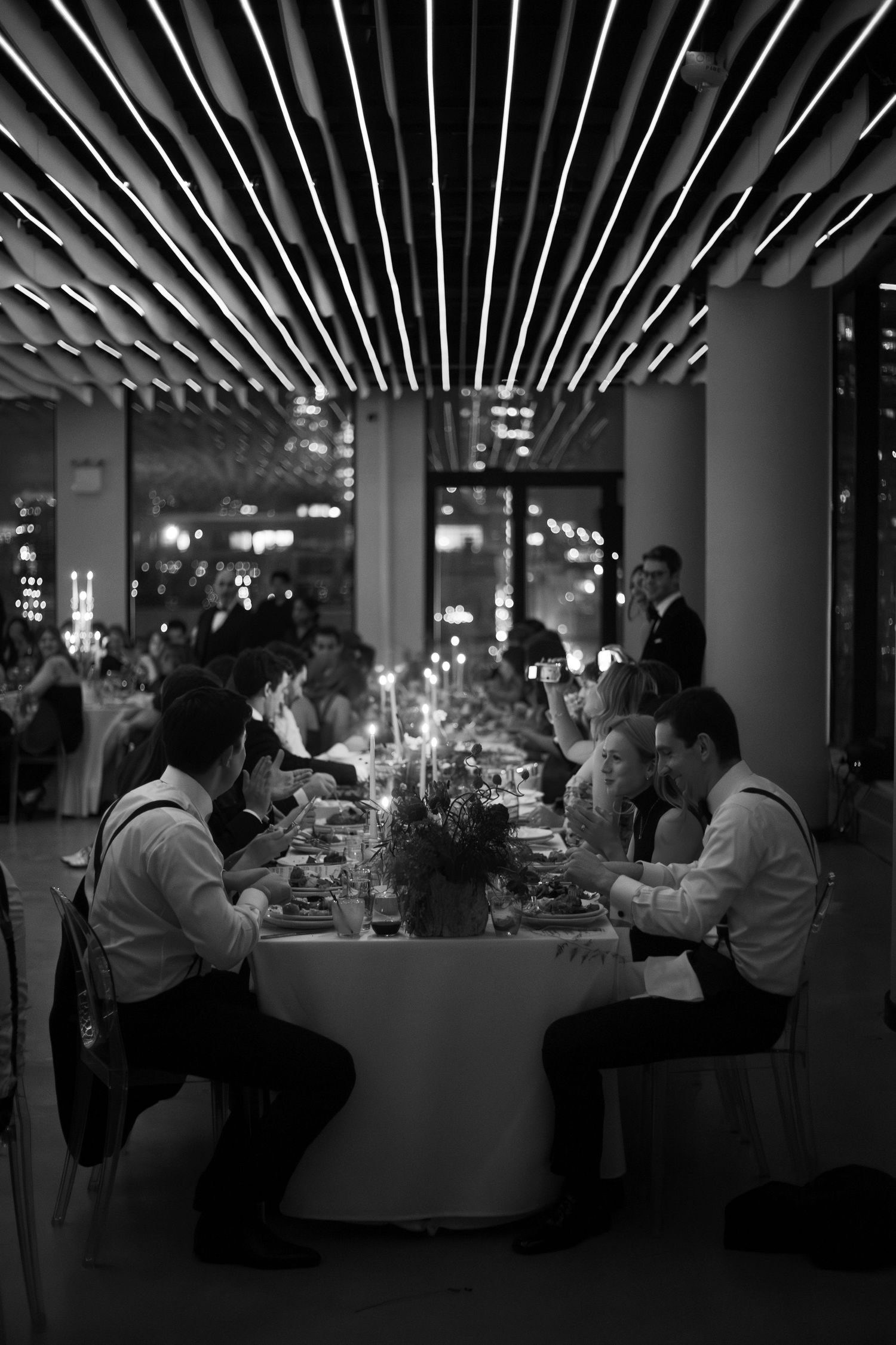 Black and white photo of elegantly dressed guests dining at a long candlelit table in a modern restaurant.