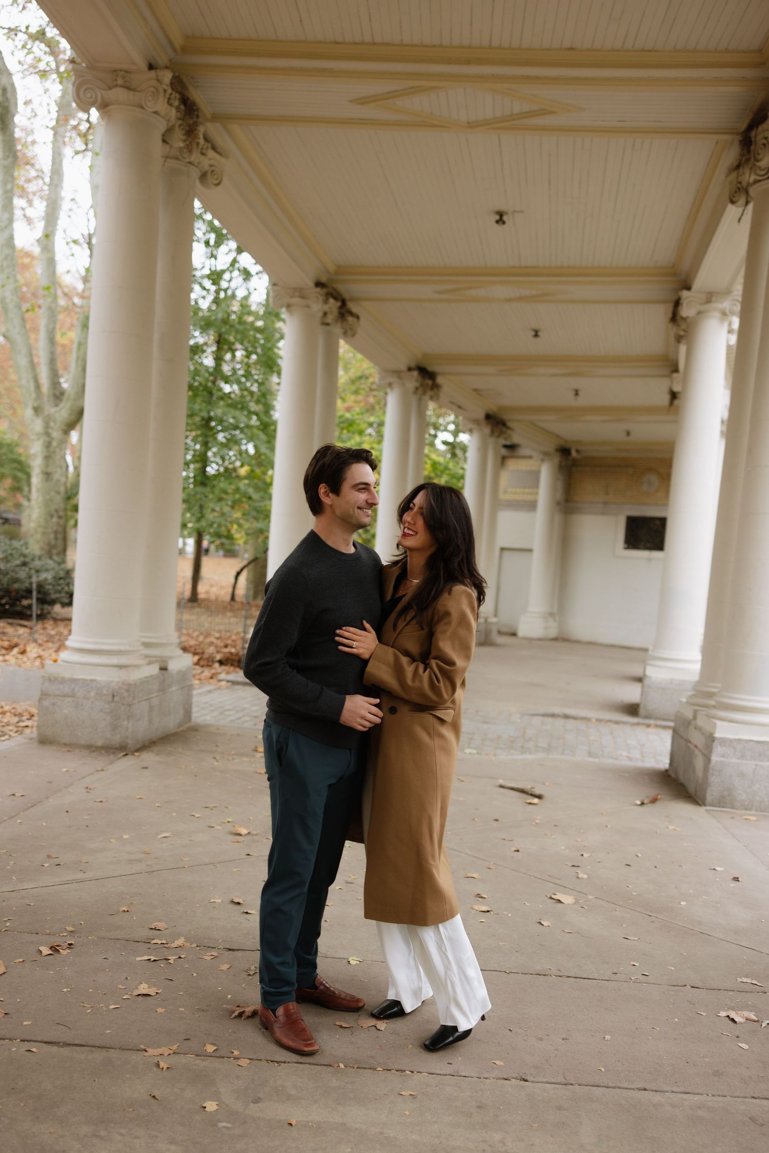 A couple shares a romantic moment under a columned walkway surrounded by autumn leaves.