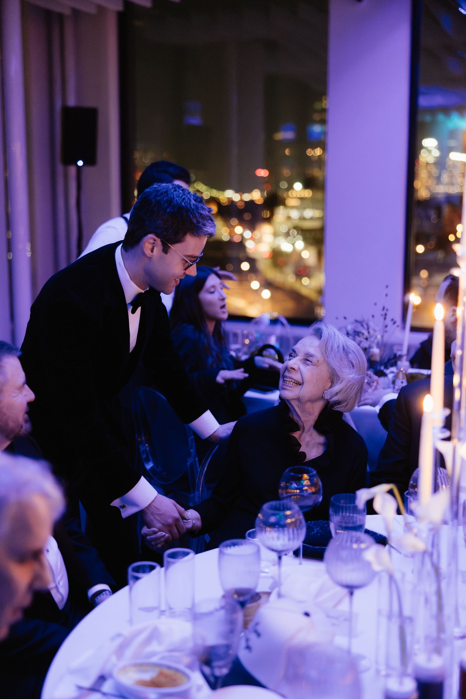 Elegantly dressed guests at a formal dinner event, with city lights visible through floor-to-ceiling windows at night.