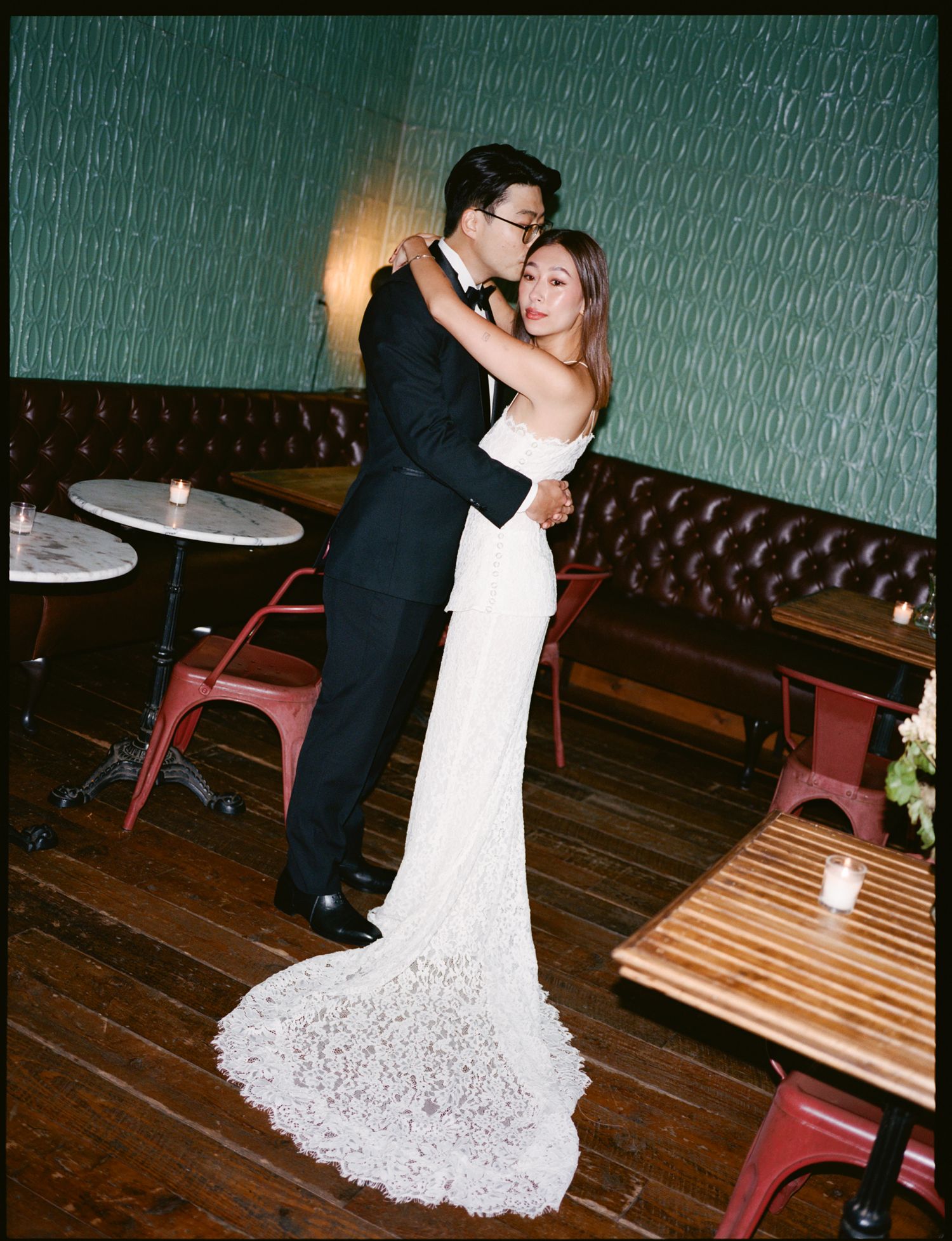 A couple in formal attire dance together in a dimly lit venue with dark wood floors and leather booth seating.