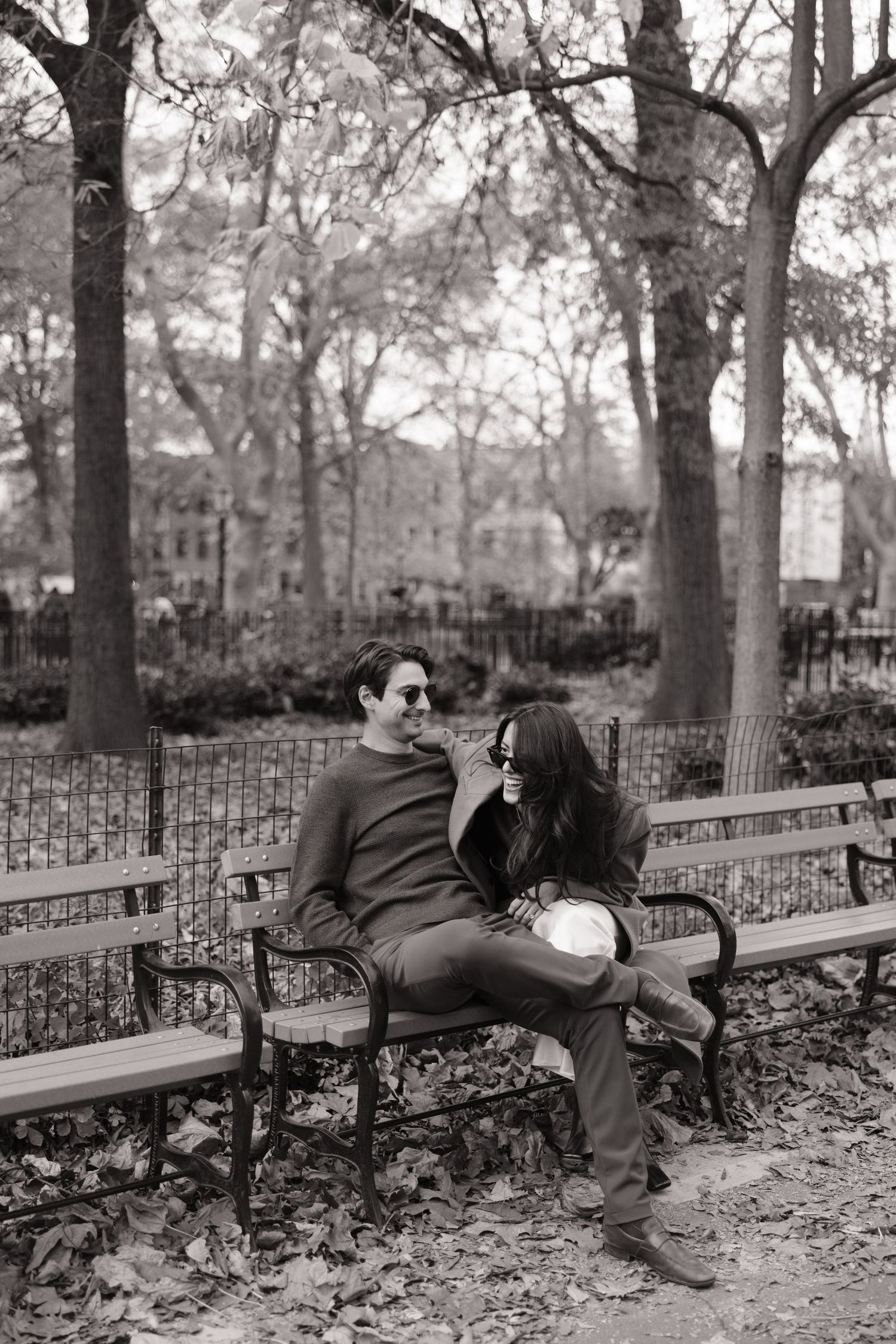 A person sleeping on a park bench in Washington Square Park, New York City, with autumn leaves scattered on the ground.