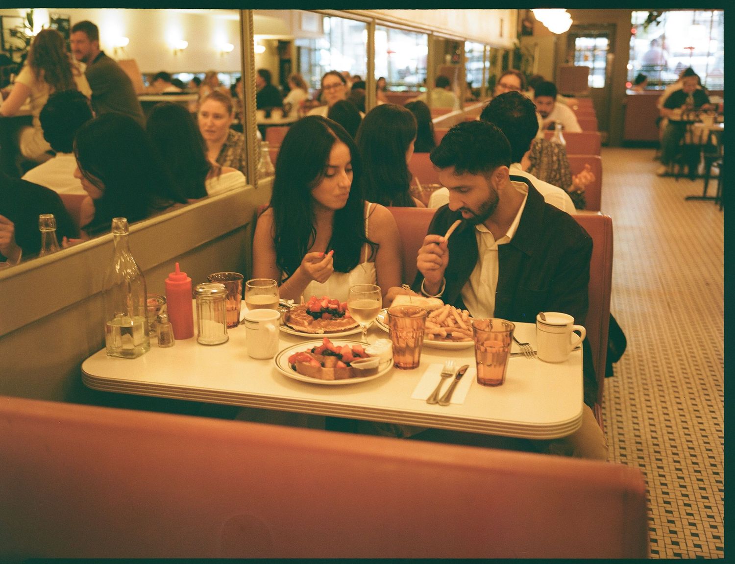 Two people share a meal at a classic American diner booth with red vinyl seats and tiled floors in warm vintage lighting.