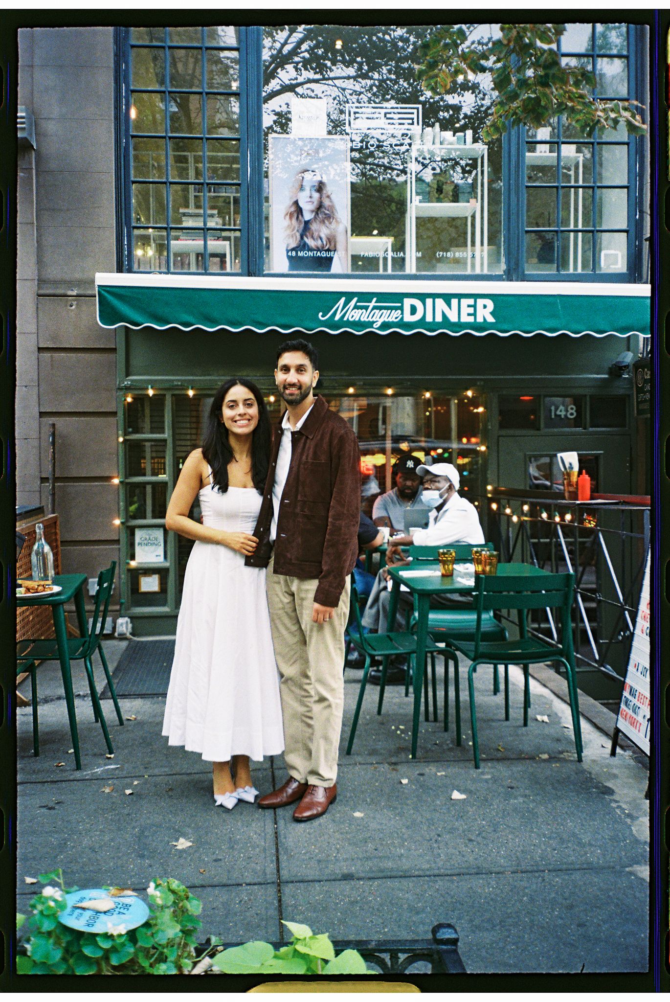 A couple stands outside a vintage-style diner with a green awning and outdoor seating on a city sidewalk.