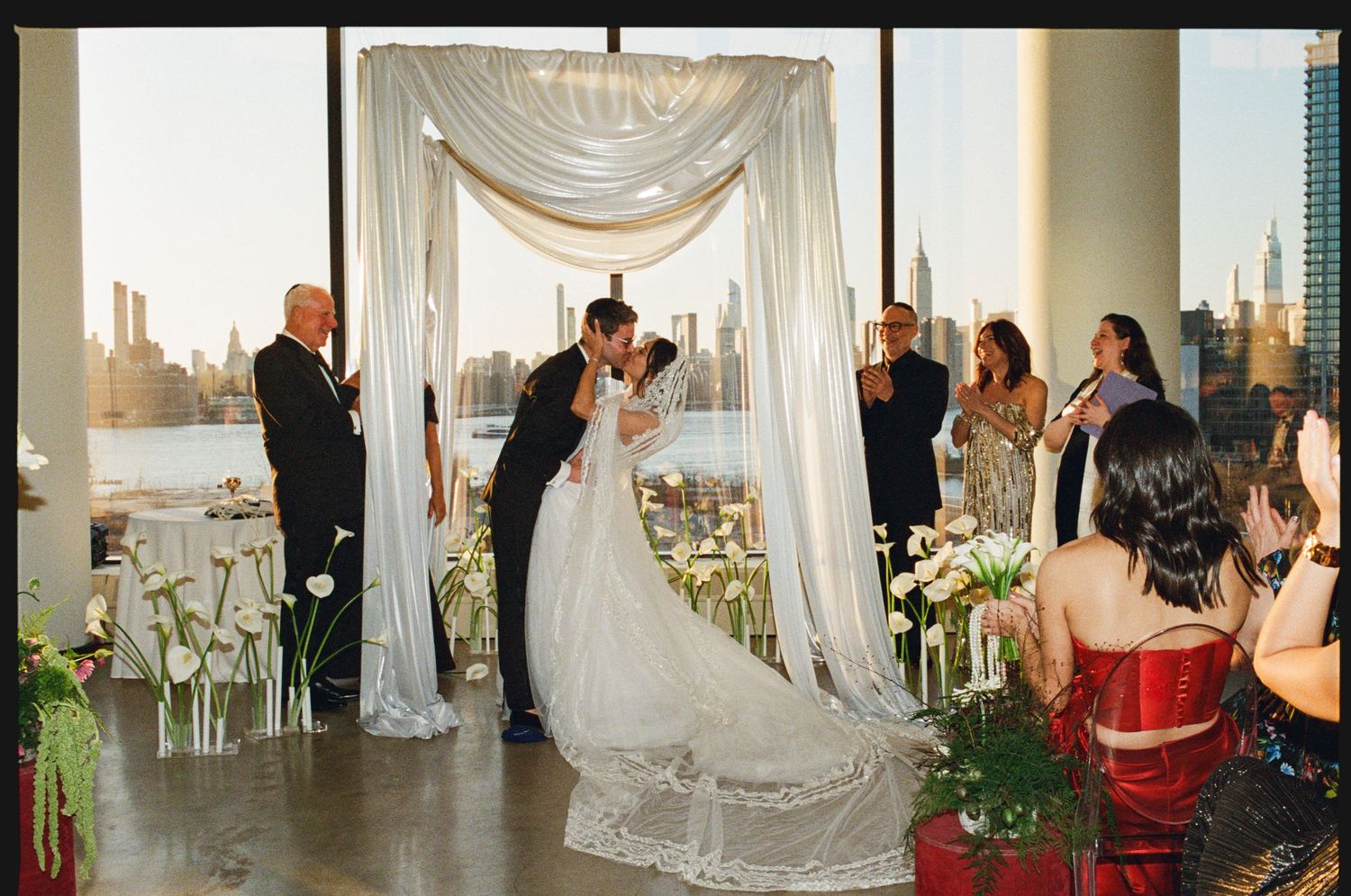 Bride and groom share first kiss at elegant waterfront wedding ceremony with NYC skyline backdrop.