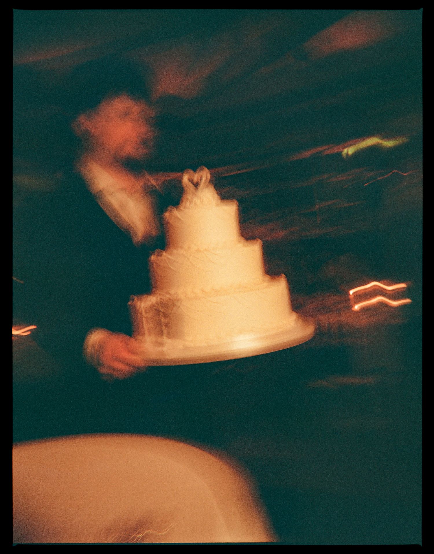 Blurry motion photo of a person carrying a white tiered wedding cake with heart topper at a dimly lit reception.