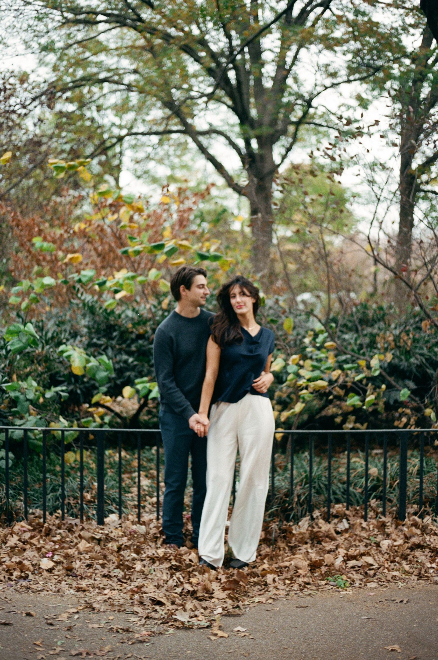A couple shares a romantic autumn moment along a park fence surrounded by fallen leaves and foliage.