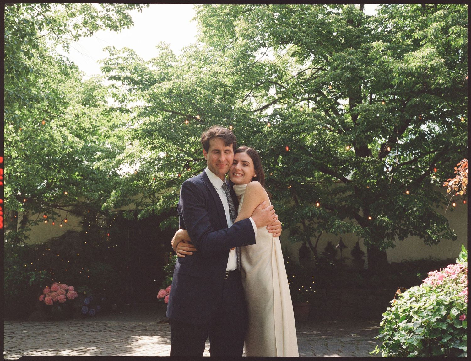 A couple in wedding attire embraces in a garden setting with lush greenery and flowering bushes in the background.