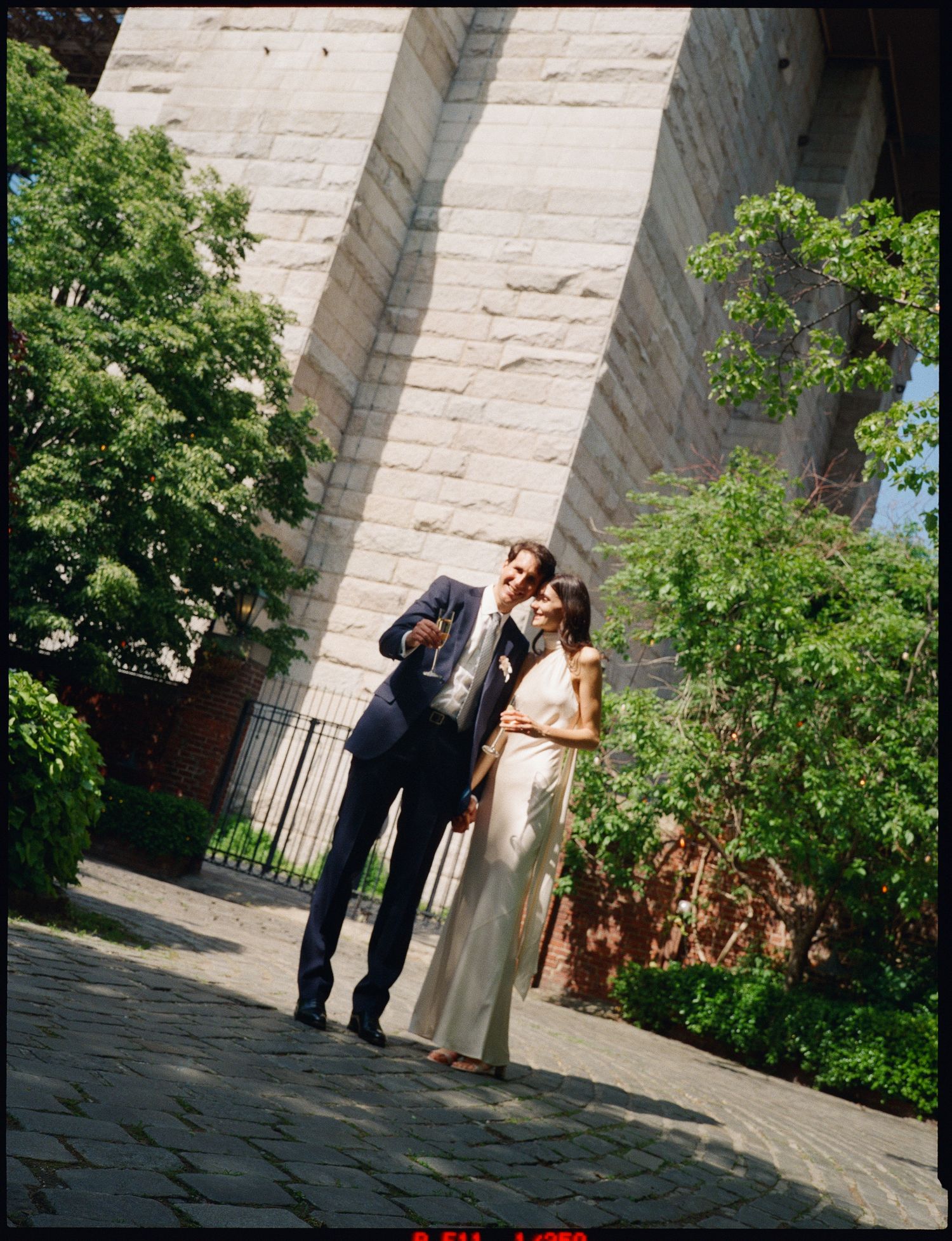 A couple shares an intimate moment in front of a stone church wall surrounded by lush greenery on a sunny day.