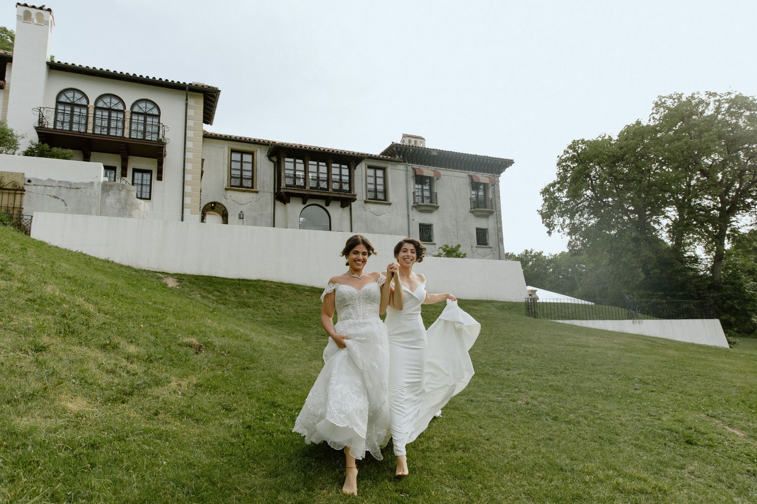 Two brides in white gowns run joyfully on a green lawn in front of an elegant historic mansion wedding venue.