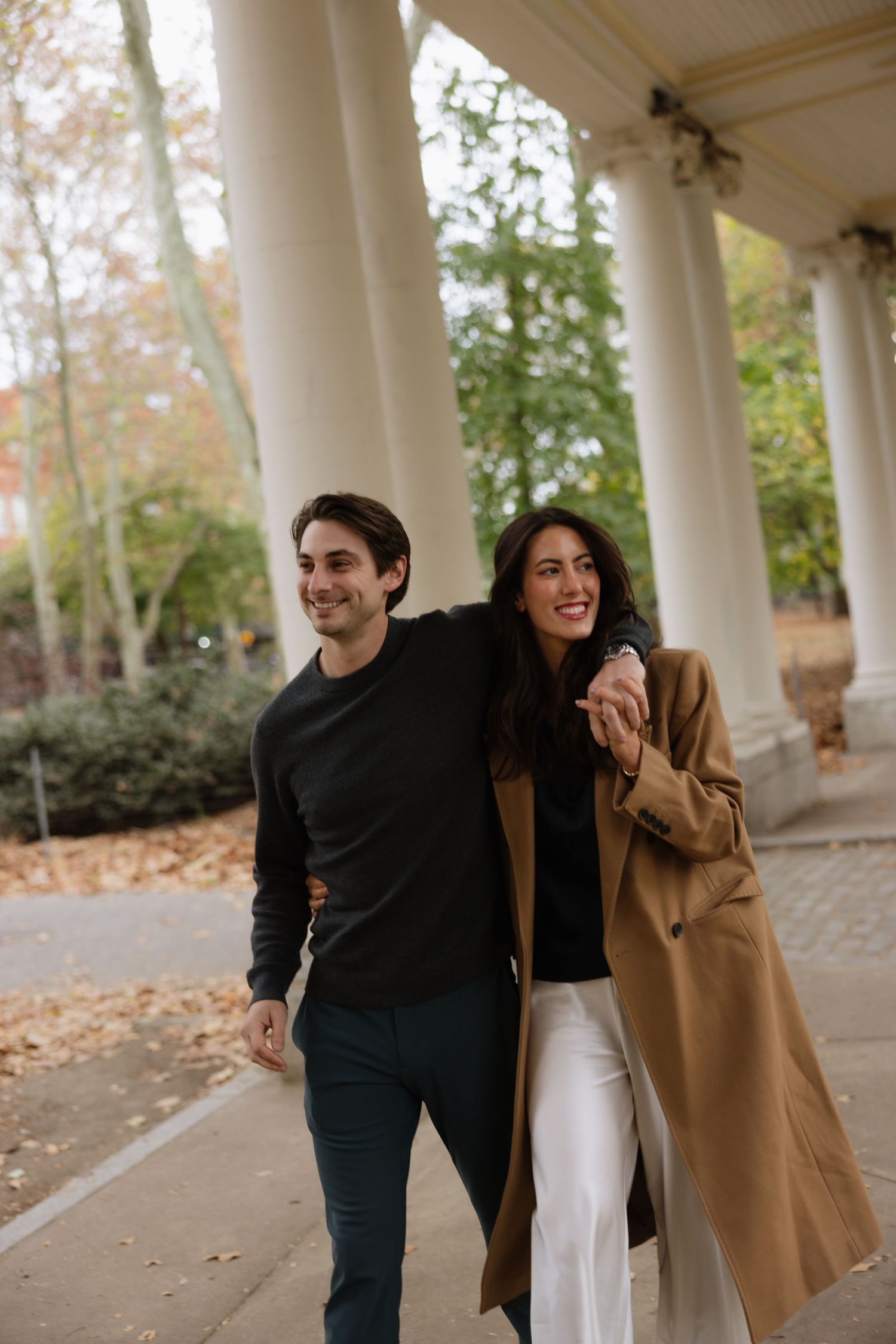 A couple in casual attire laughs together while walking under white columns on an autumn day.